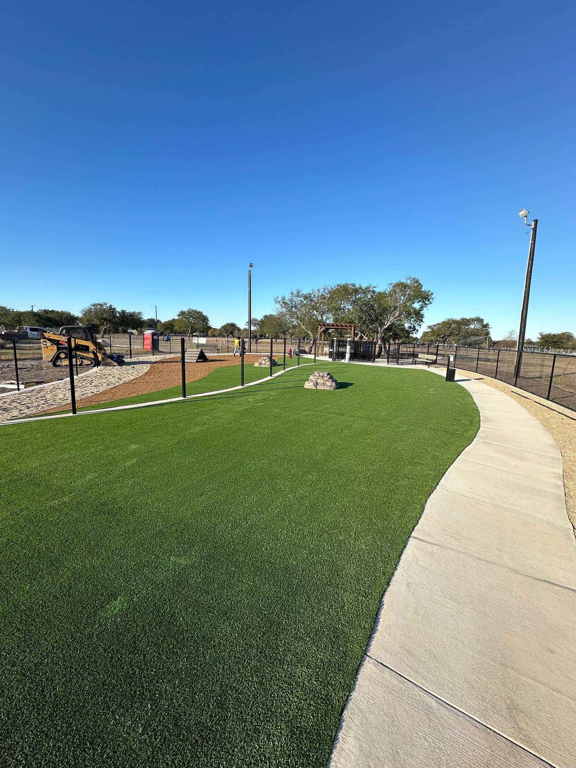 Curved sidewalk beside a green lawn under a clear blue sky, with a landscaped park and trees in the distance.