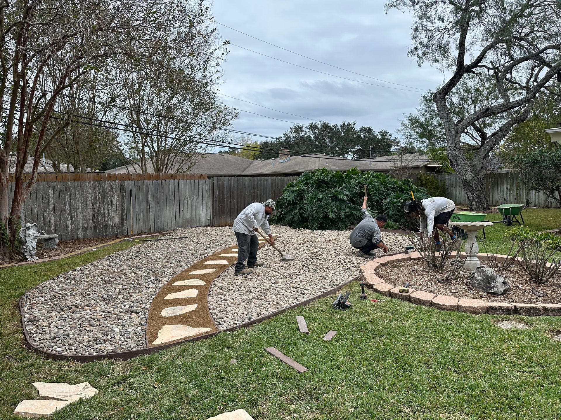 Three people work in a suburban backyard, leveling gravel around a curved stone pathway and a circular garden bed.