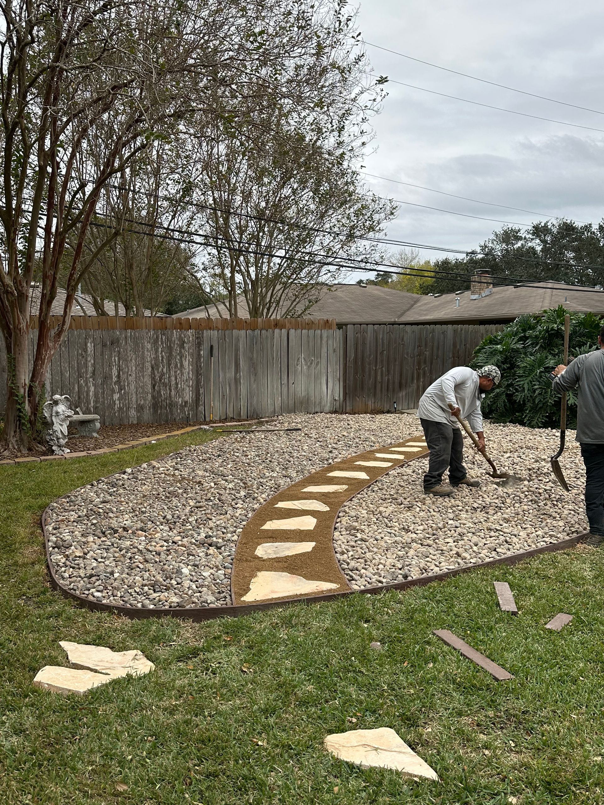 Two people landscaping a backyard, installing stone pavers into a gravel bed bordered by edging.