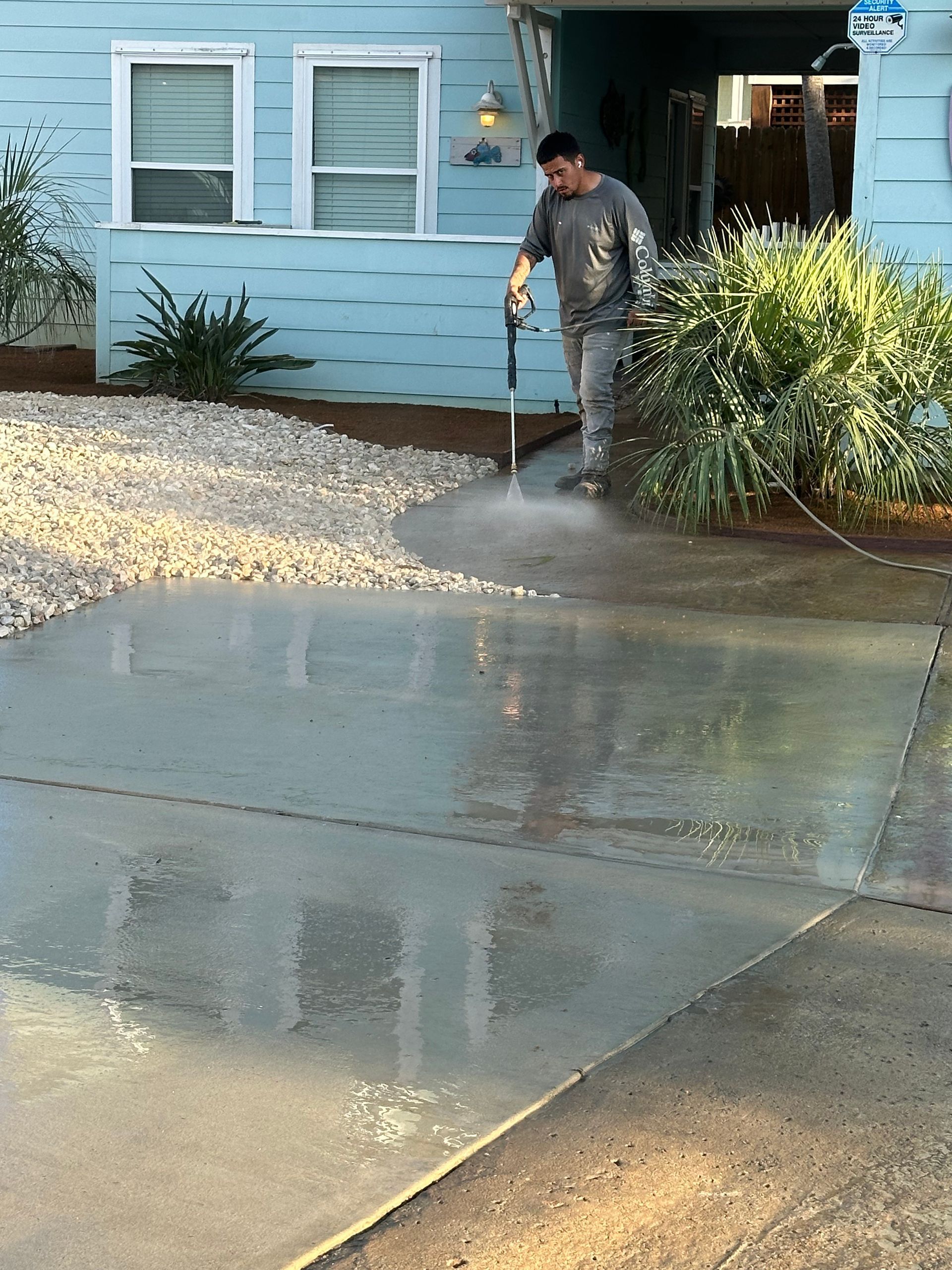 A person uses a pressure washer to clean a concrete driveway in front of a blue house.