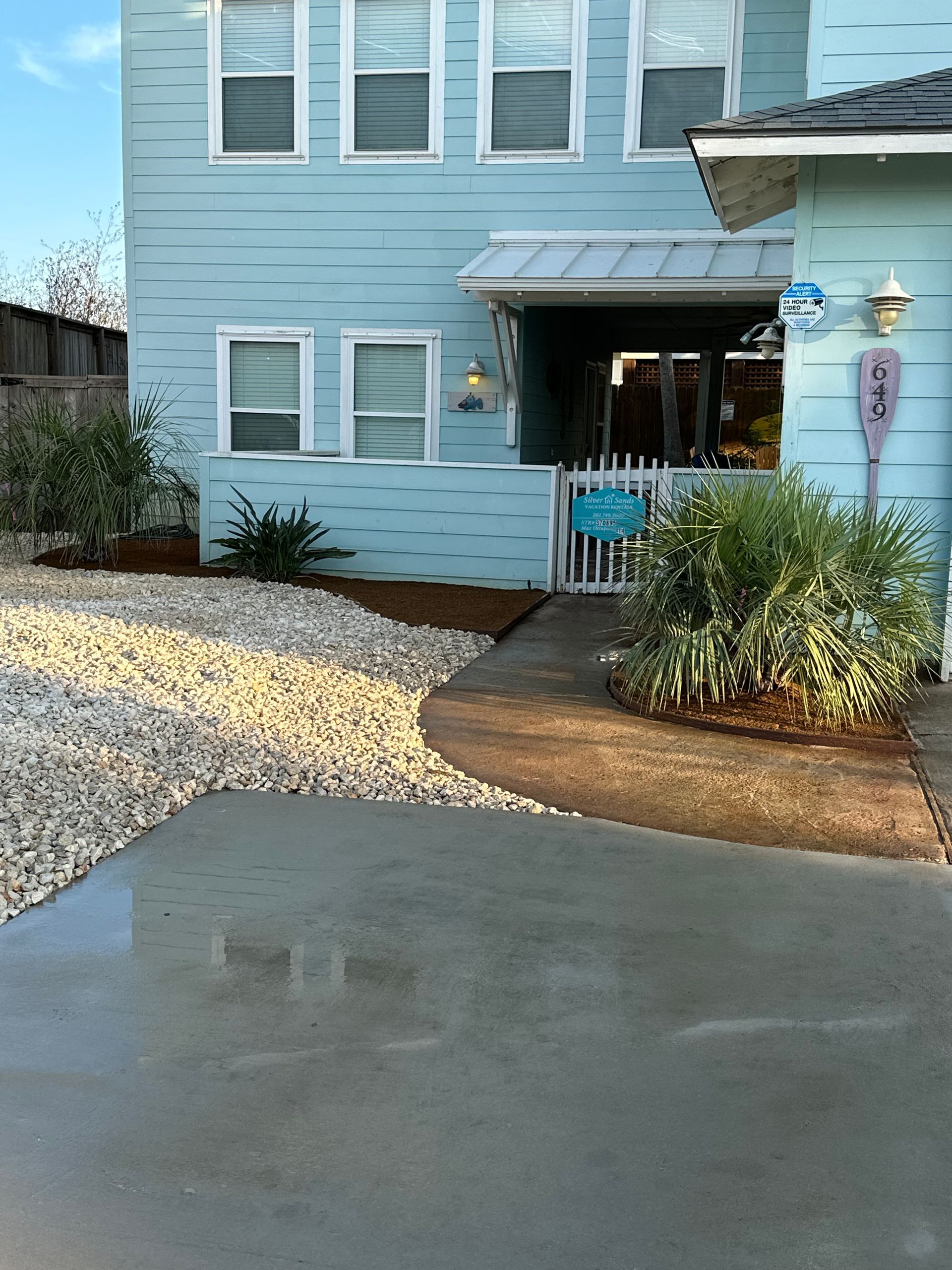 A light blue house exterior with white trim, a concrete walkway, gravel landscaping, and a small gated entrance.