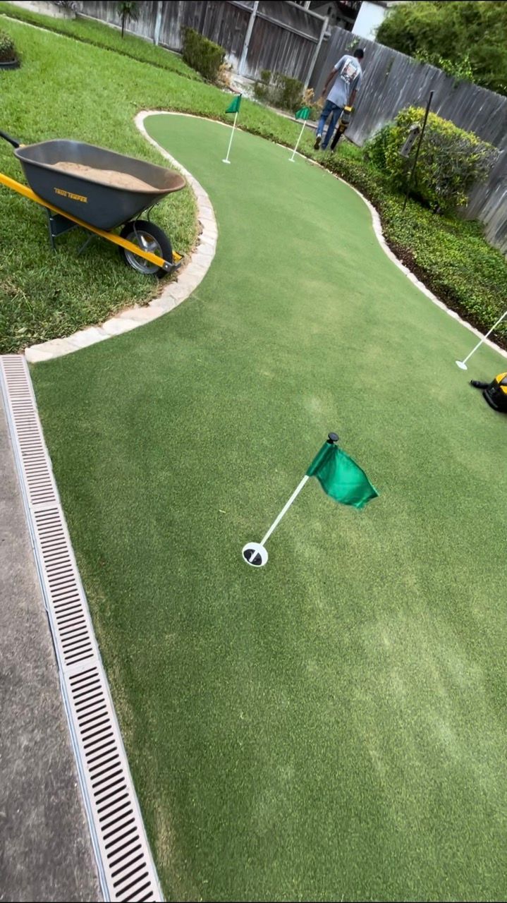 A person works on a backyard putting green with artificial grass, golf flags, and a wheelbarrow nearby.