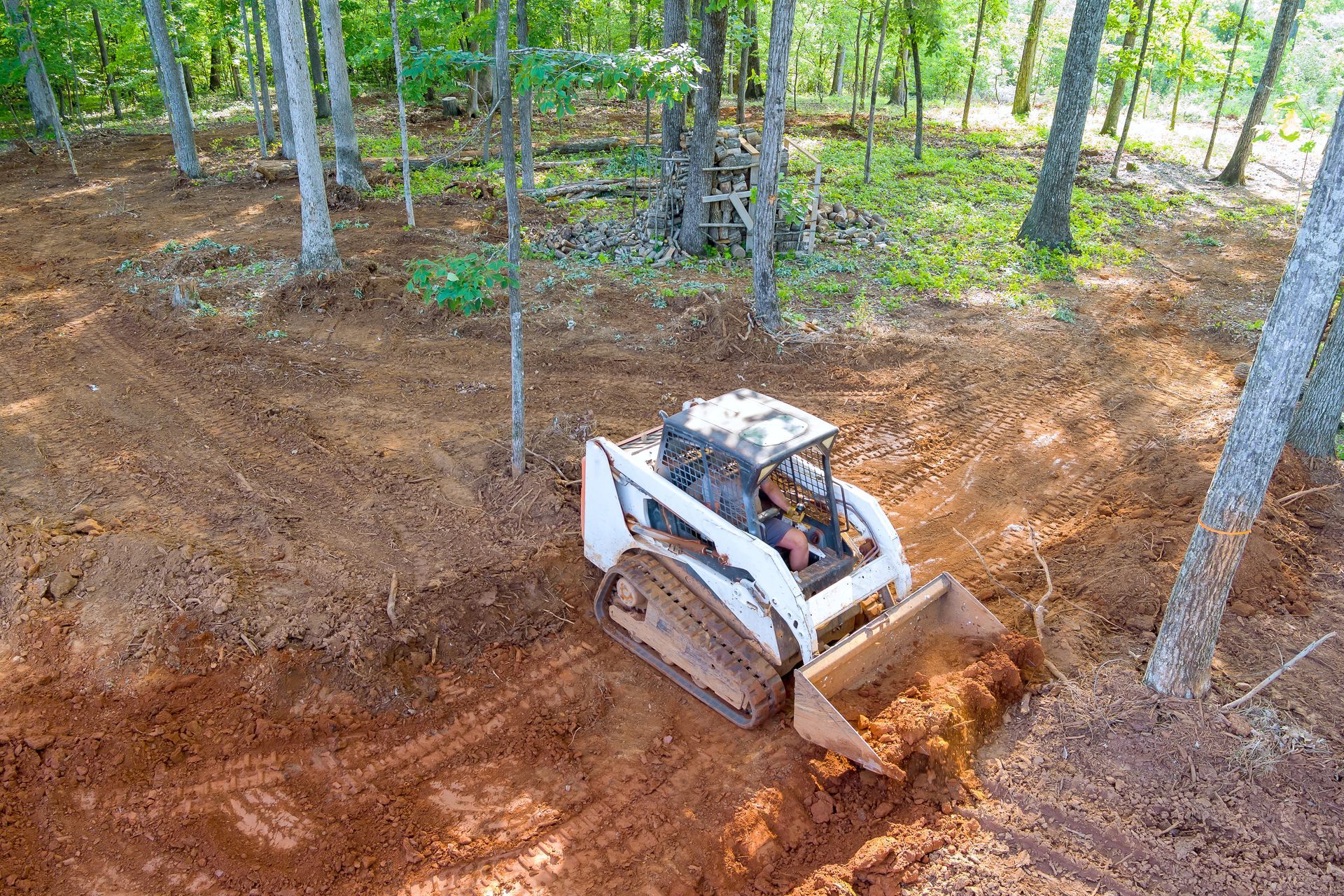White skid steer pushes dirt in a forest clearing with trees around it.