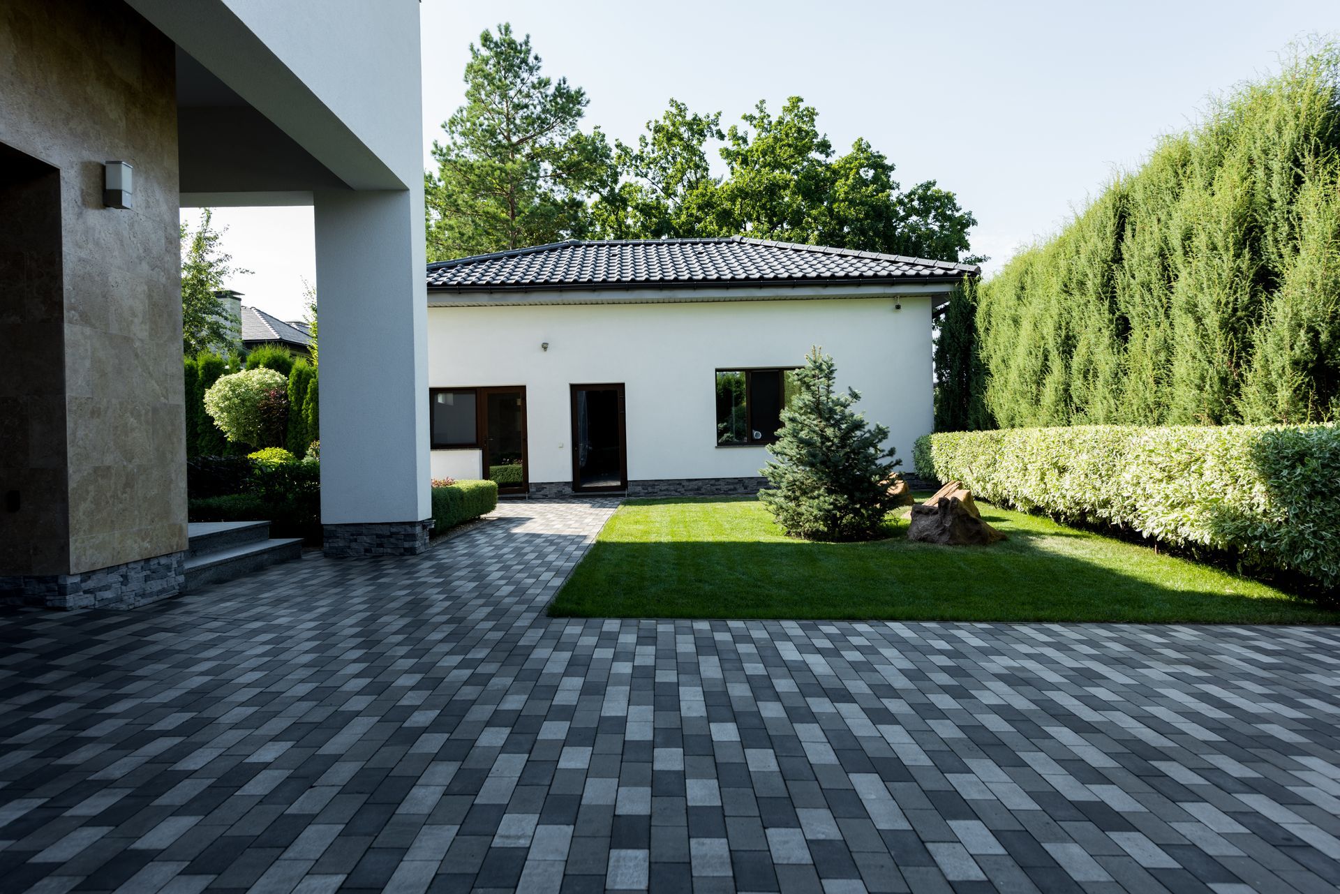 Modern house courtyard with gray pavers, green lawn, and tall hedges in bright sunlight