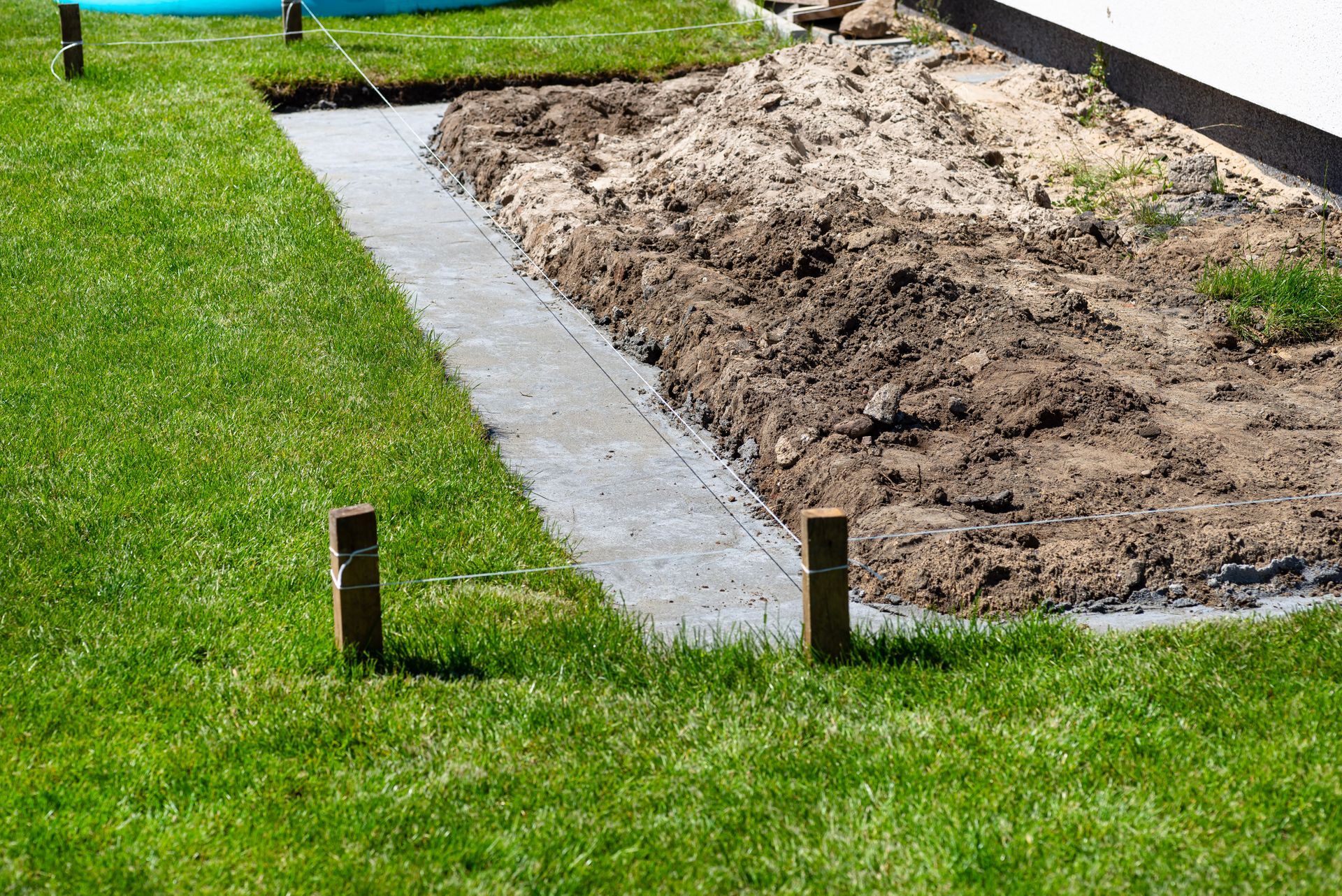 Partially dug yard beside a concrete path, with piles of dirt and green grass around it