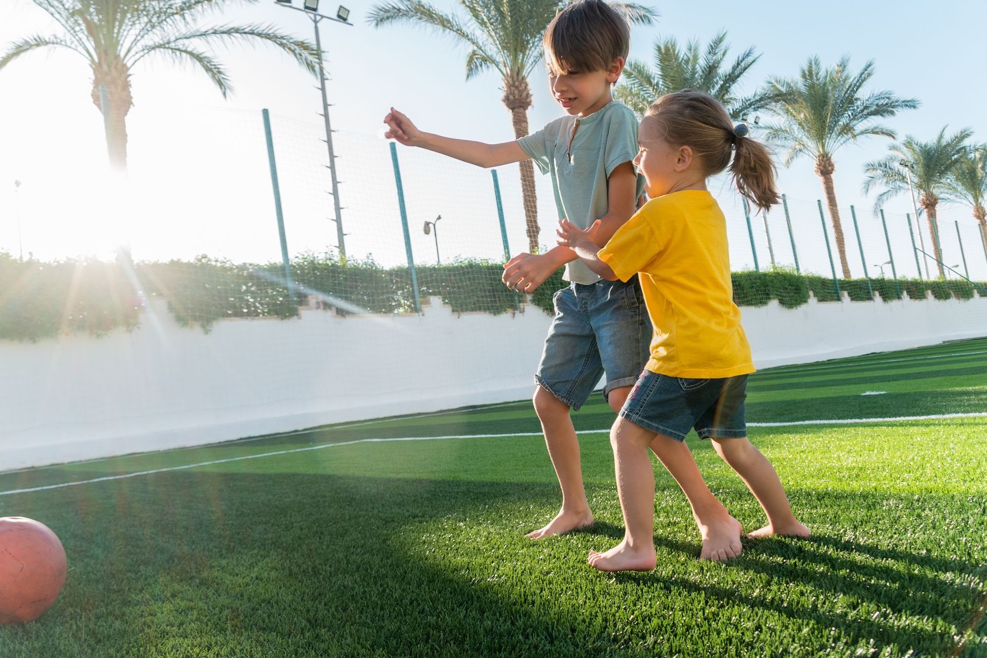 Two children playing with a red ball on a grassy field with palm trees in the background under bright sunlight.