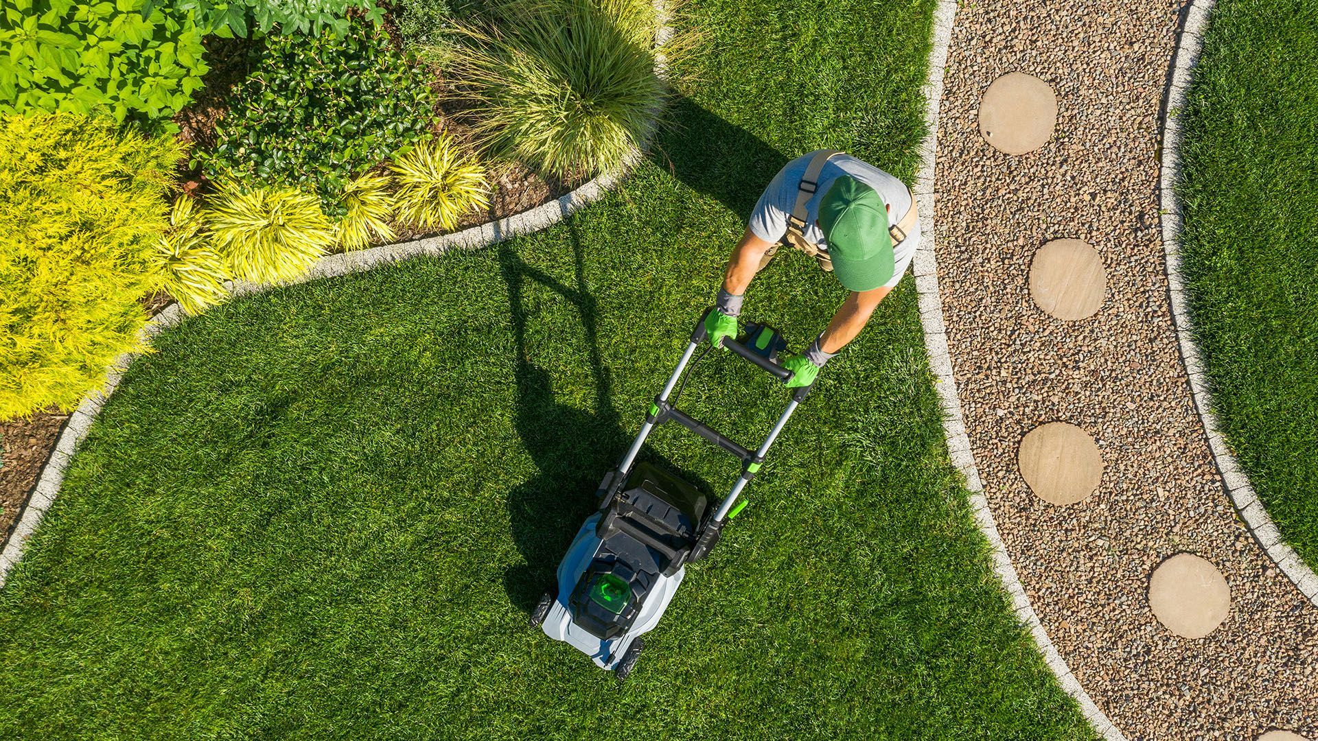 Person mowing a lush green lawn with a mower, near a pebble path and flowerbeds.