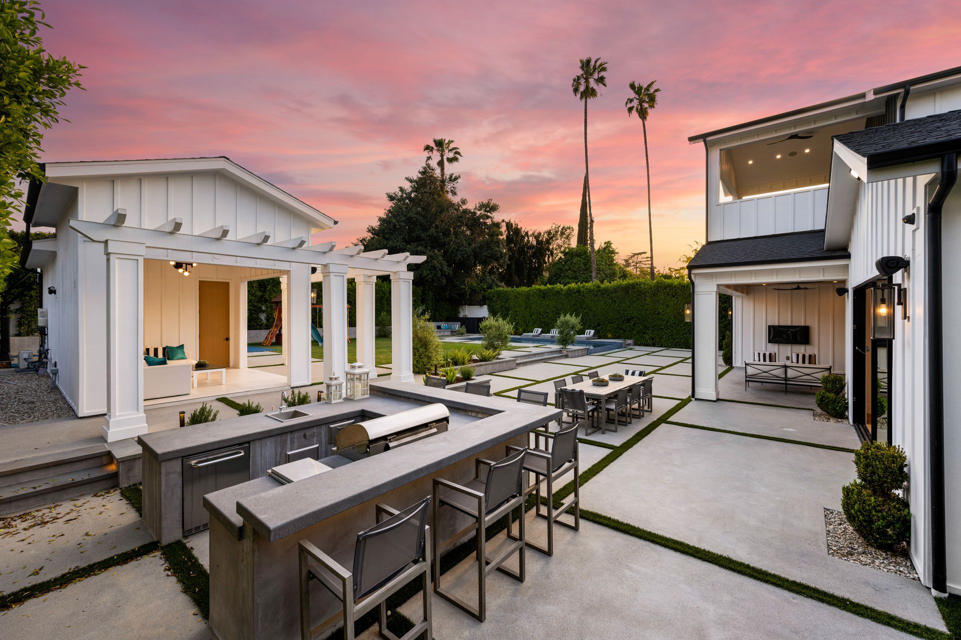 Modern backyard patio with white pergolas, fire pit seating, and a pink sunset sky