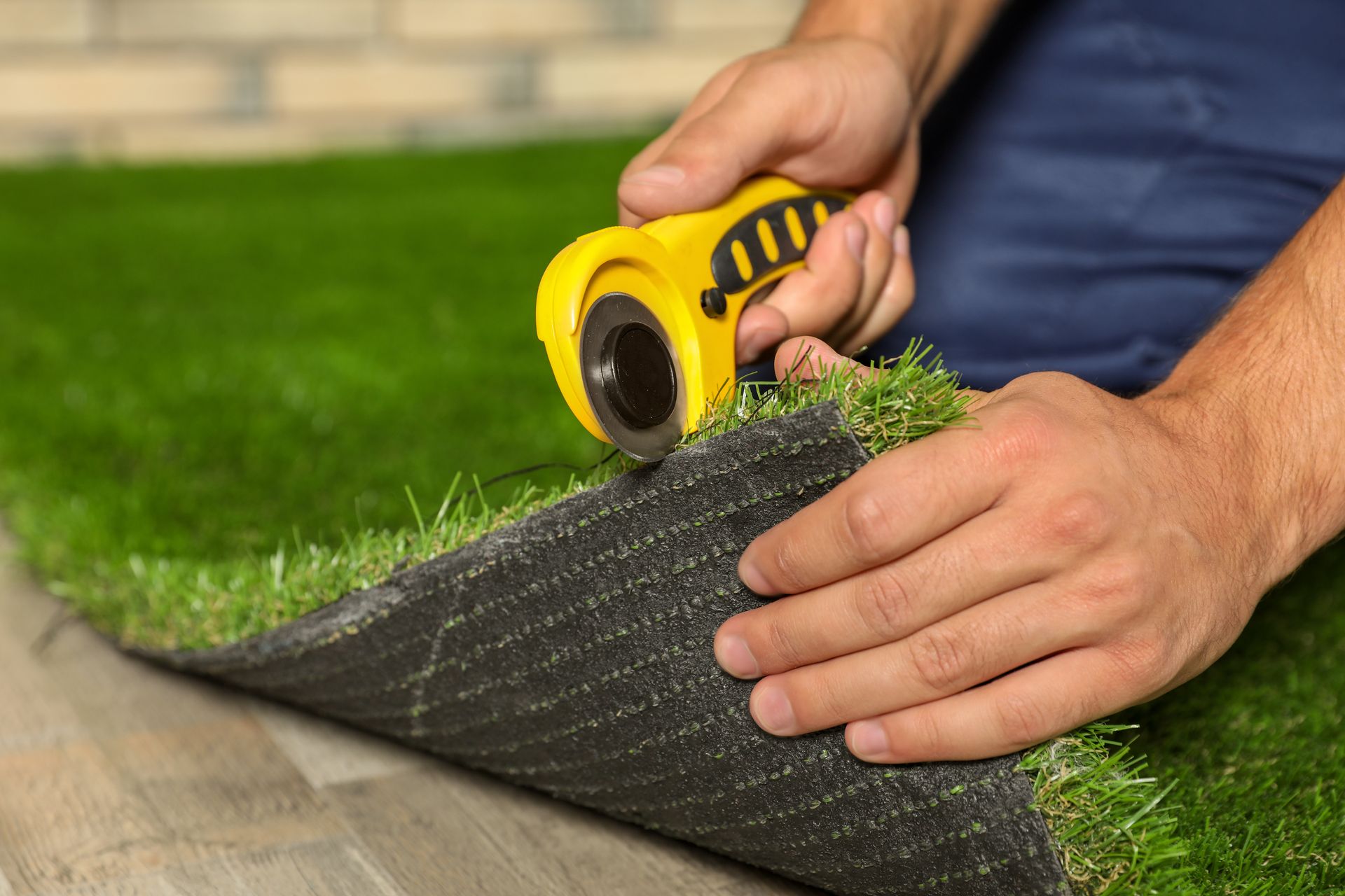 Hands use a yellow rotary cutter to trim the edge of a piece of artificial grass on a floor.
