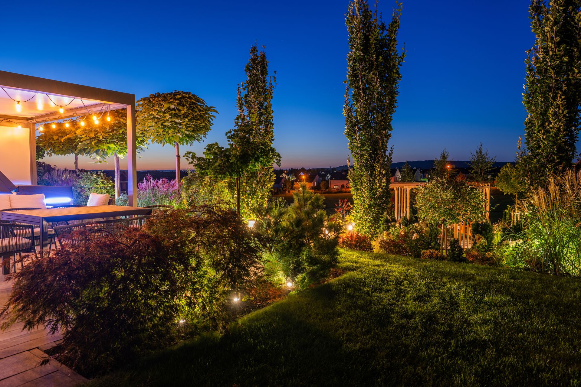 Illuminated garden patio at dusk with a covered seating area, tall trees, and colorful landscape lighting