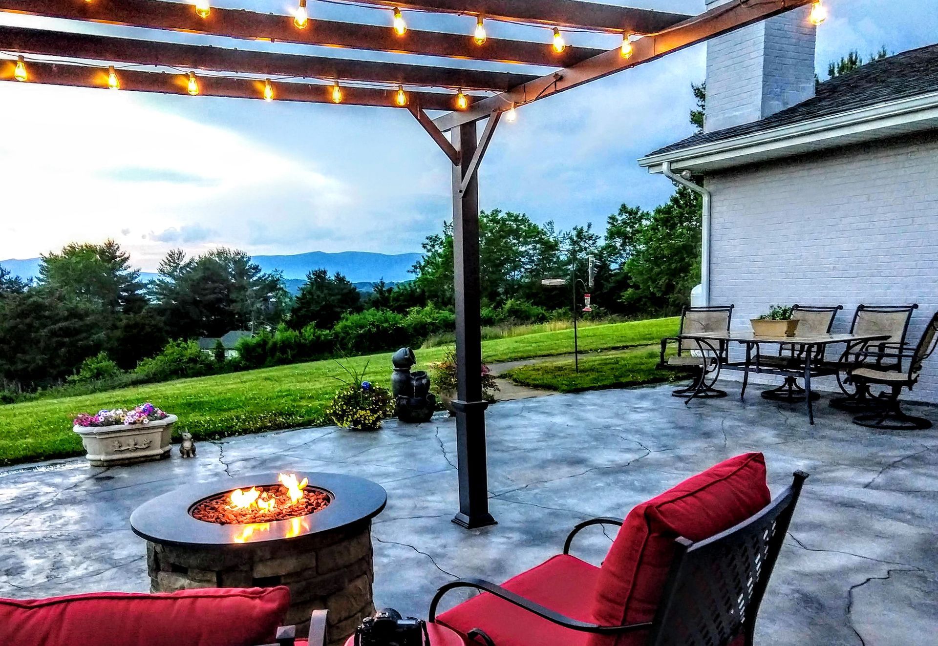 Covered patio with string lights, fire pit, red chairs, and a lawn view at sunset