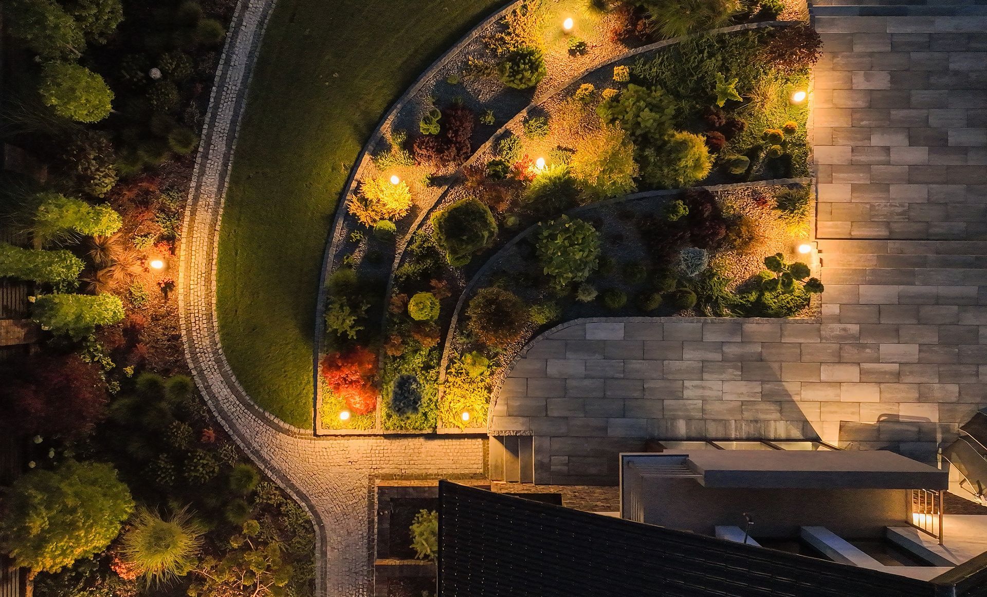 Overhead view of a curved garden path with glowing lights, greenery, and adjacent stone paving at night