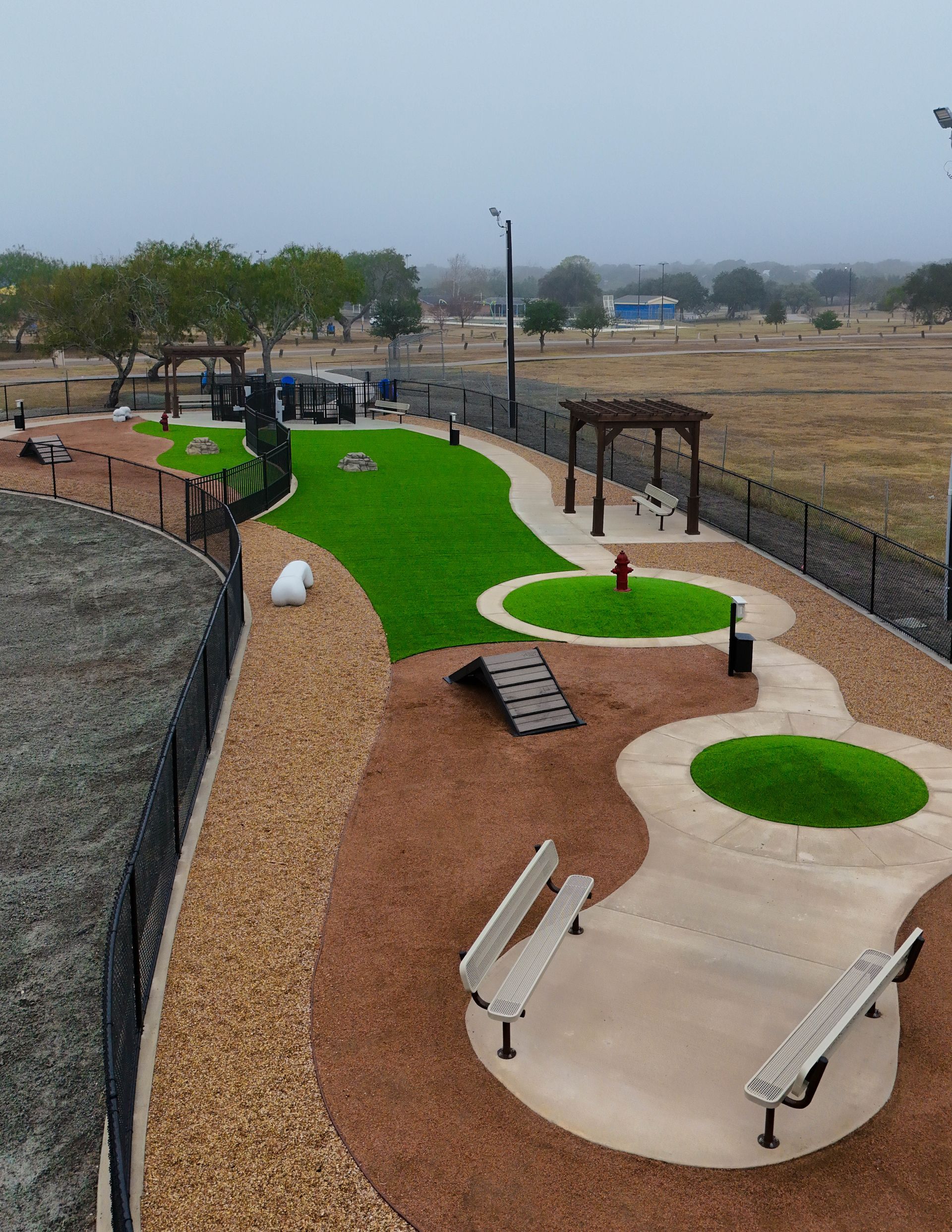 A high-angle view of a fenced dog park featuring artificial grass, gravel paths, agility ramps, seating, and a pergola.
