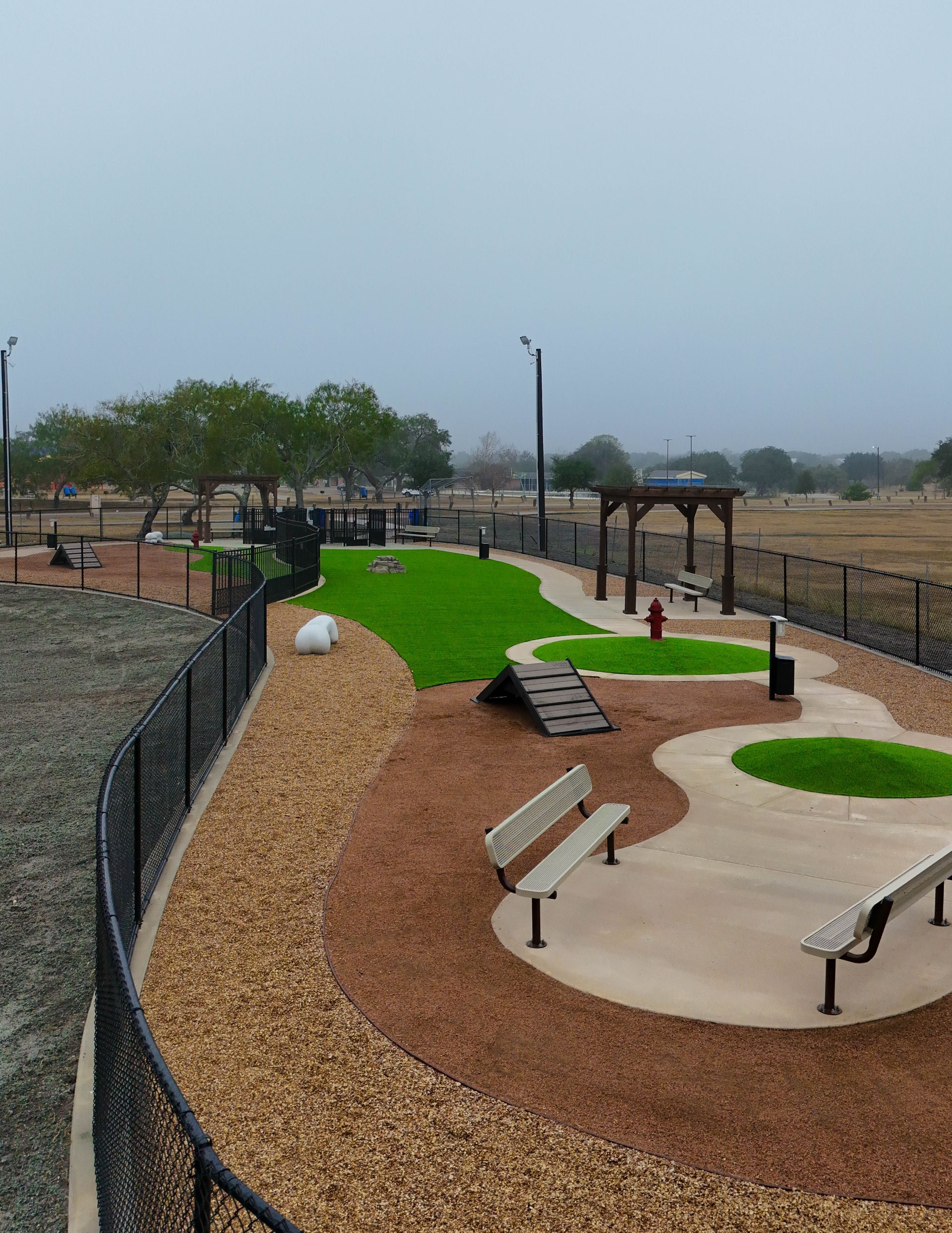 An outdoor fenced dog park with agility ramps, artificial grass patches, benches, and a shaded structure.