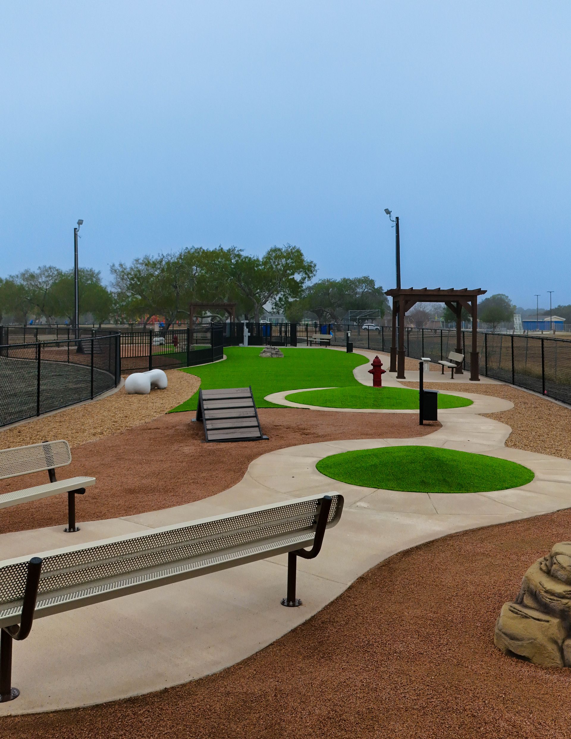 A fenced dog park with green turf areas, a wooden ramp, a red fire hydrant, and benches along a winding concrete path.