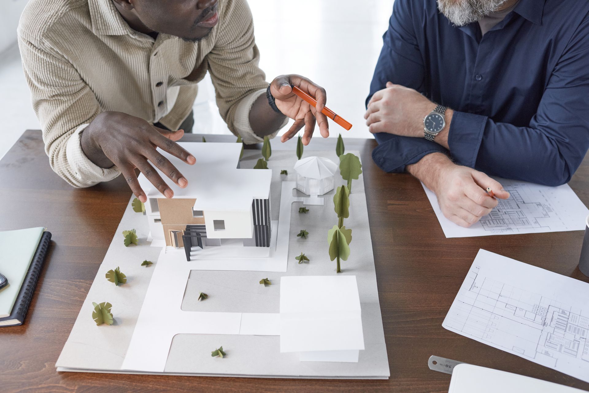 Two men are sitting at a table looking at a model of a house.