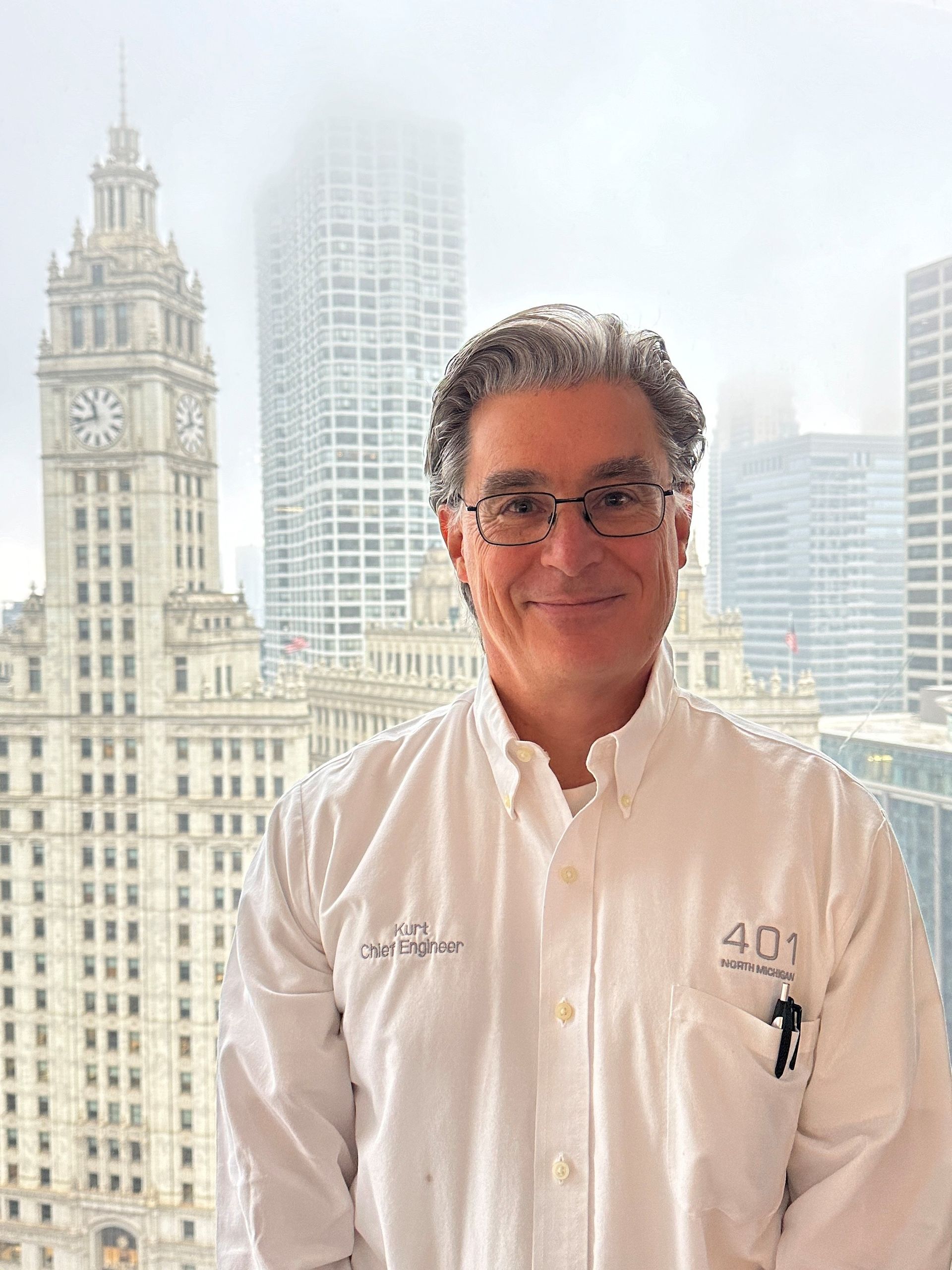 A man in a white shirt is standing in front of a window with a city skyline in the background.