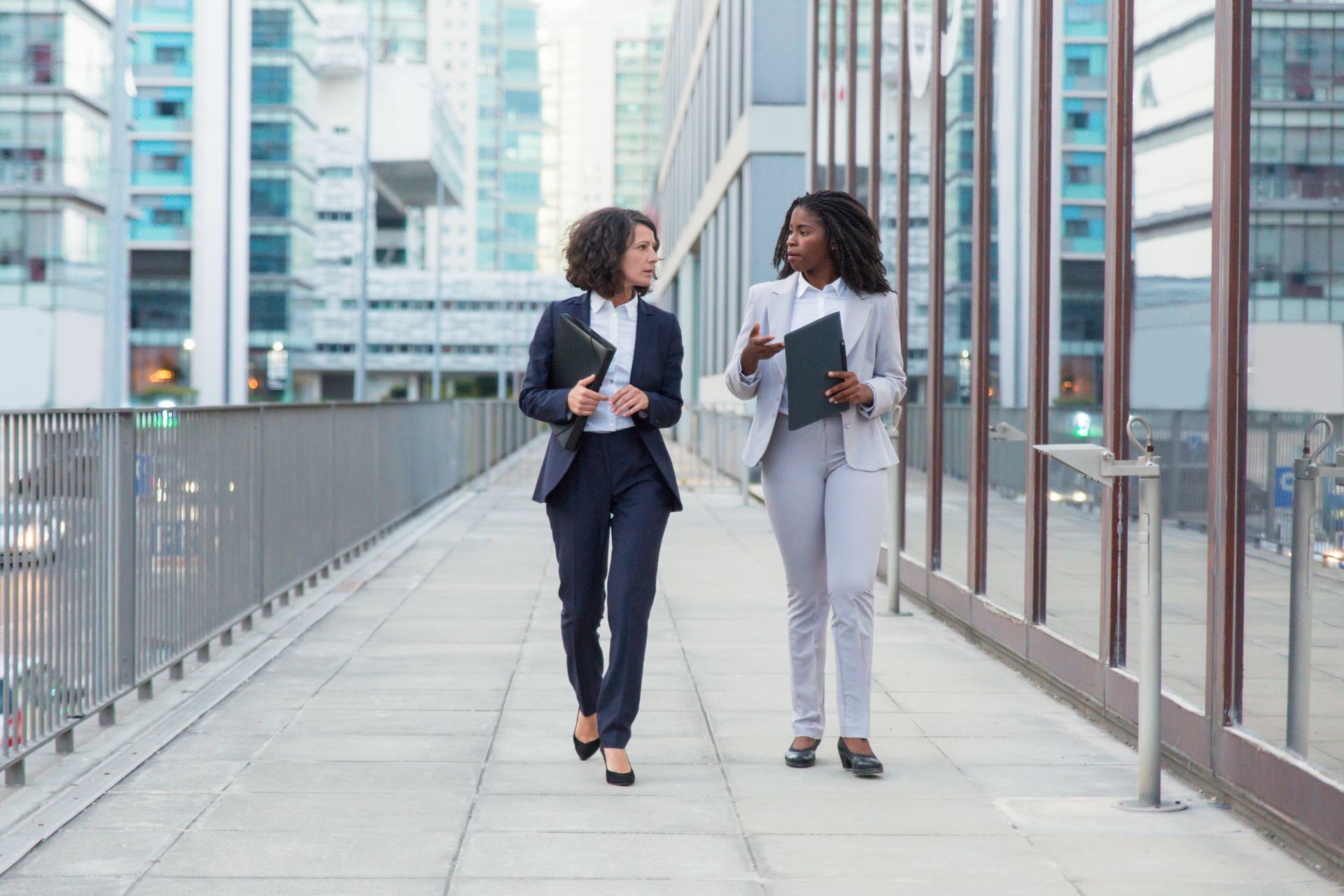 Two business women are walking down a sidewalk in front of a building.
