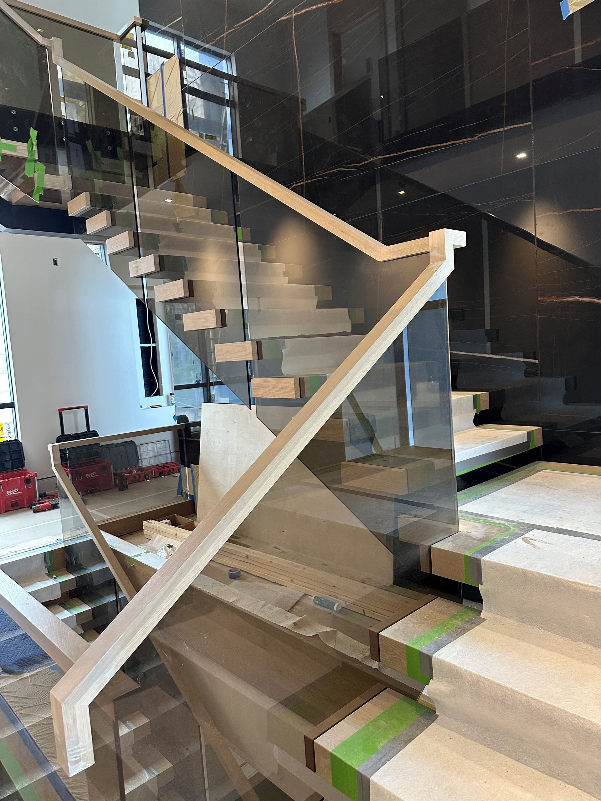 Staircase under construction with glass panels and wooden handrails. Beige steps and dark wall in the background.