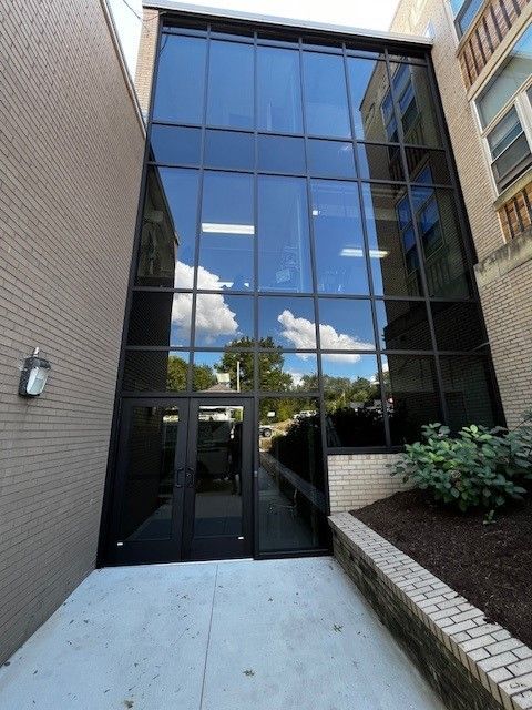 Glass-walled entrance between brick buildings. Double doors lead to a walkway. Sky and trees reflect in the windows.