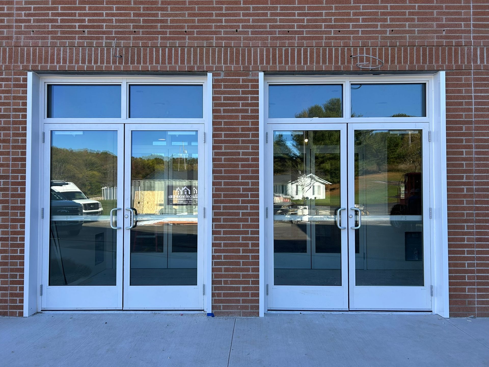 Two white-framed glass doors set in a red brick building; reflections show vehicles and other buildings.