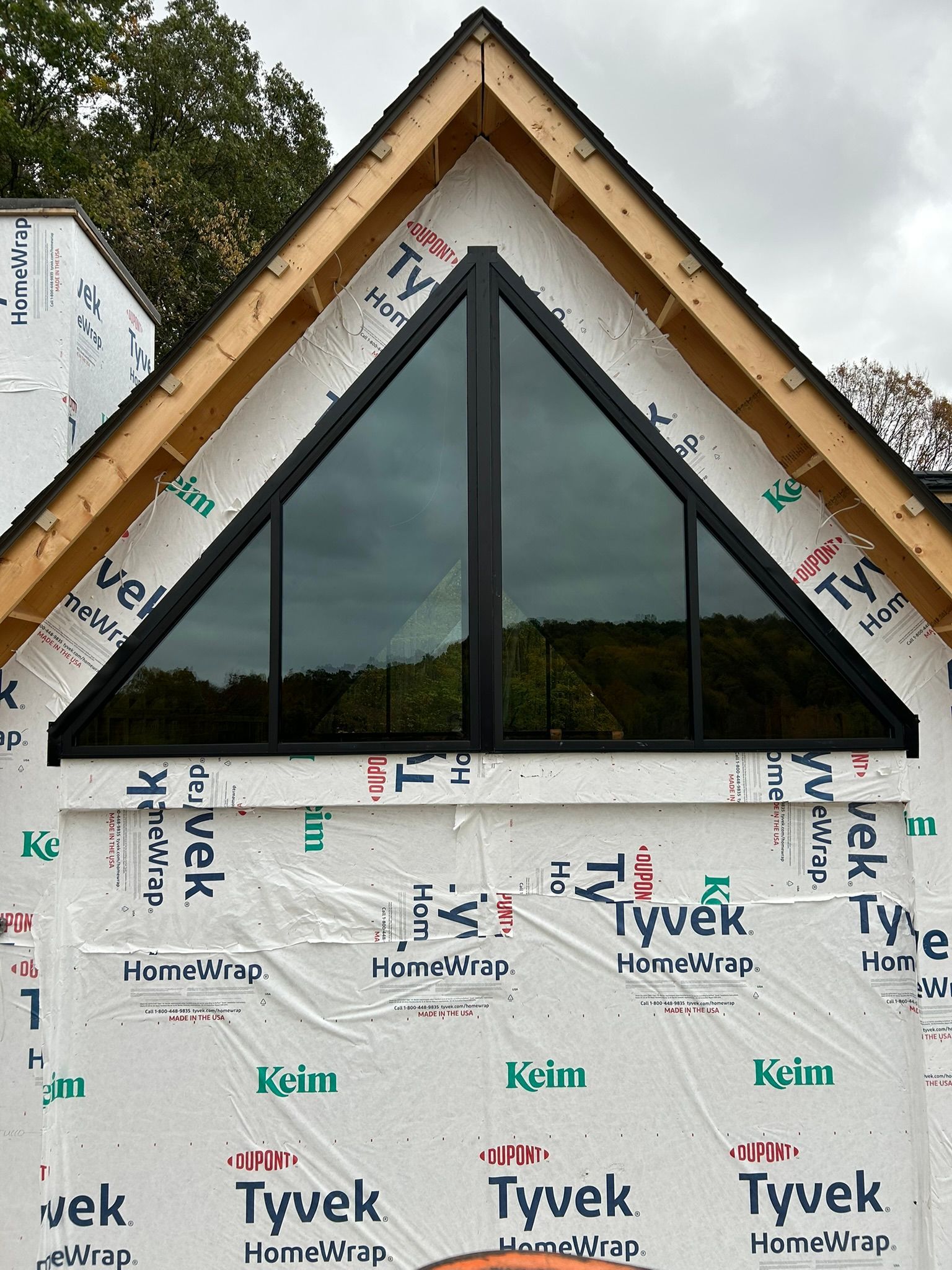 Triangular black-framed window installed in a house gable. The house is under construction, covered in Tyvek wrap.