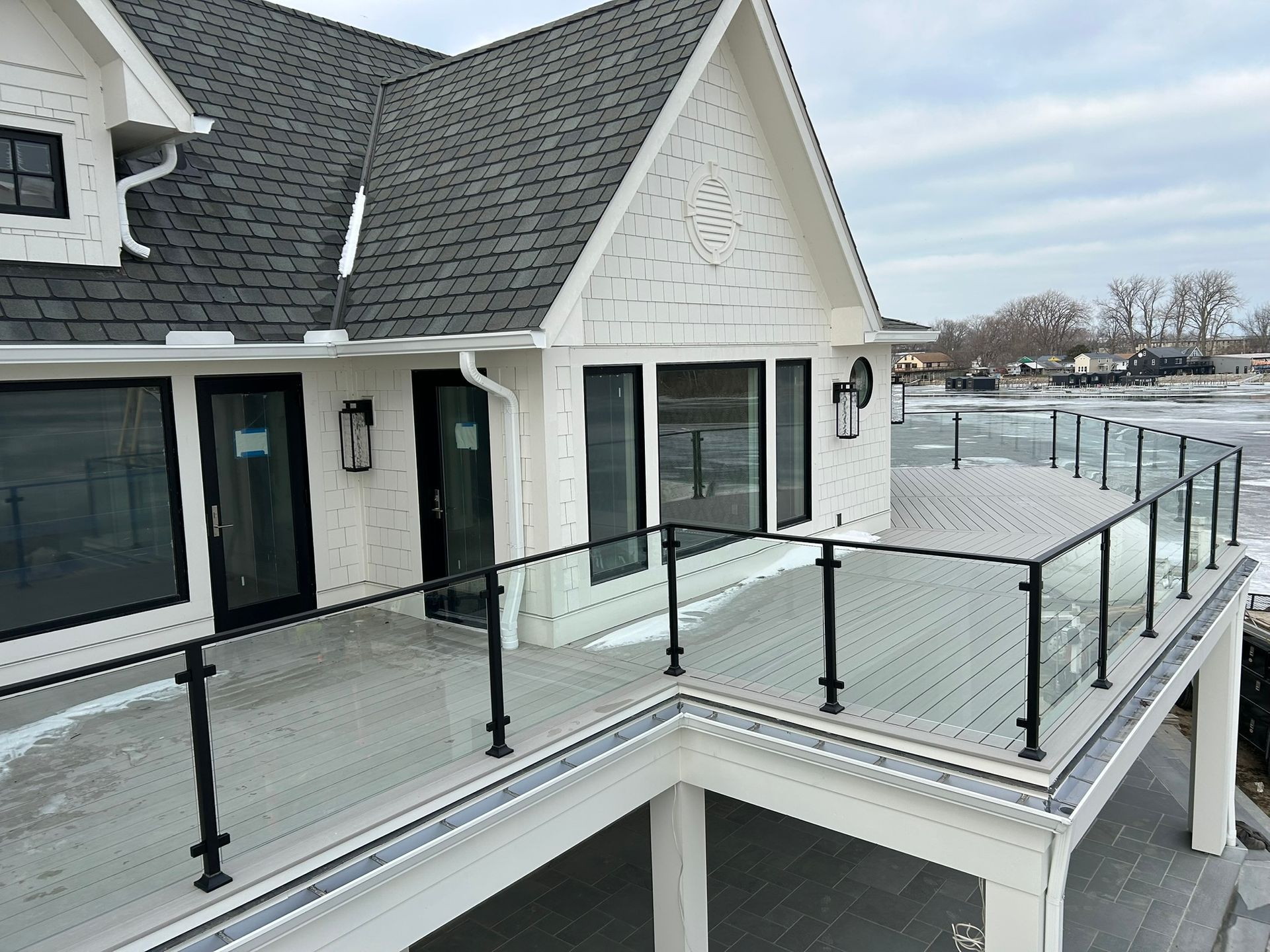 White house with black-framed windows and a glass-railed deck overlooking water. Overcast day.