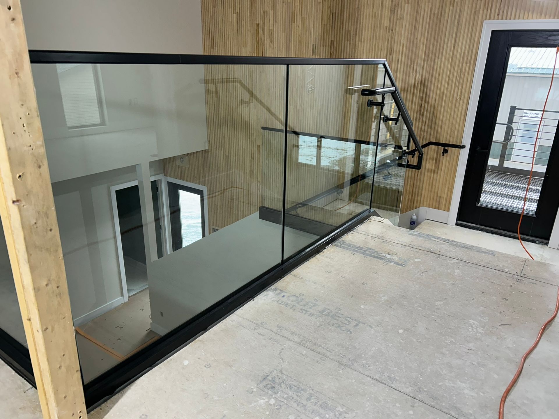 Interior view: Glass railing on a staircase with a black frame; wood paneling on the wall, door in the background.