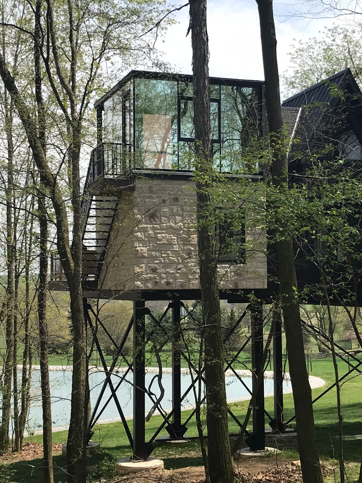 Modern glass-walled treehouse on metal supports, surrounded by trees, near a lake.