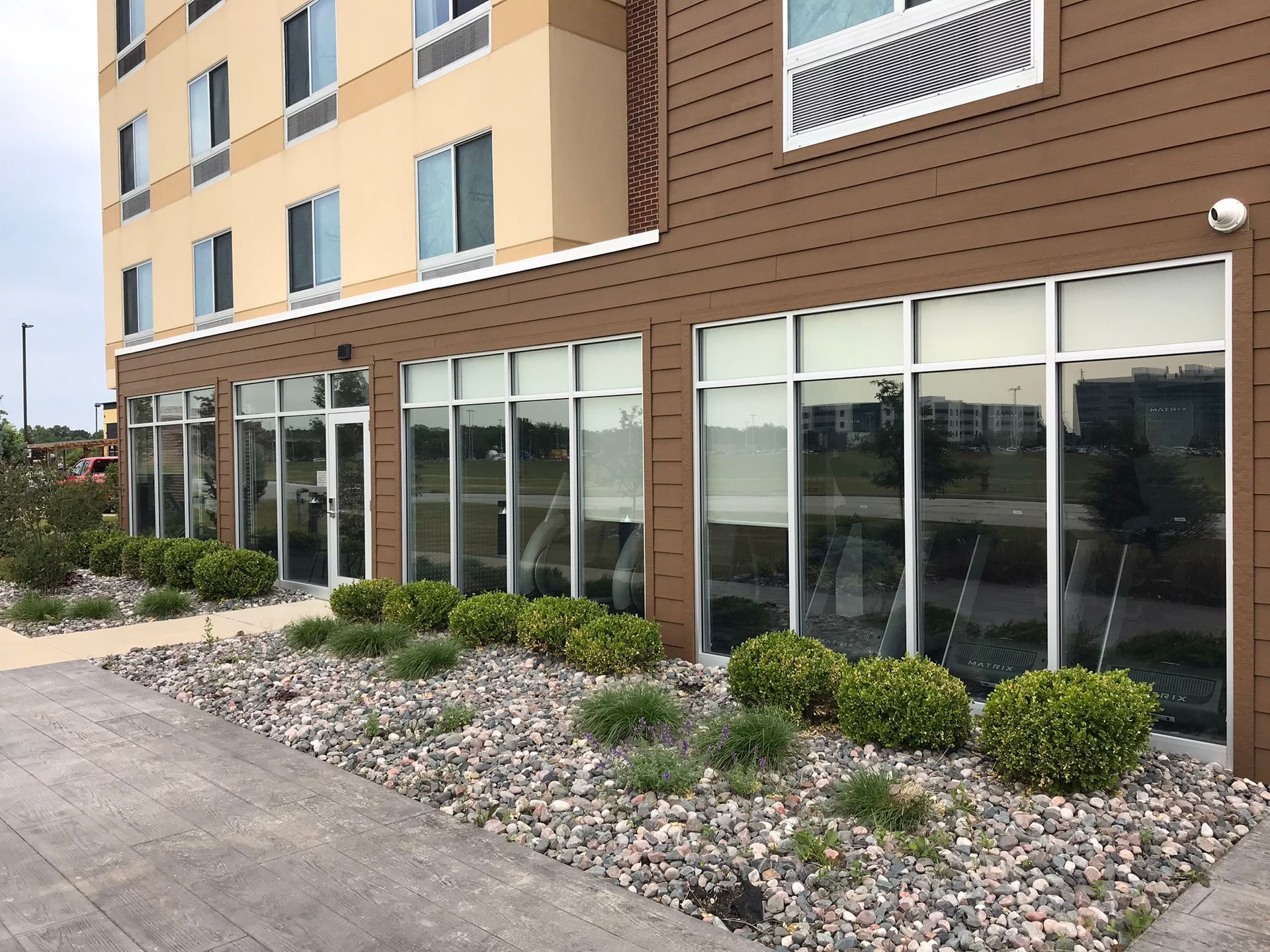 A modern hotel with large windows, tan and brown facade, and landscaping with bushes and rocks.