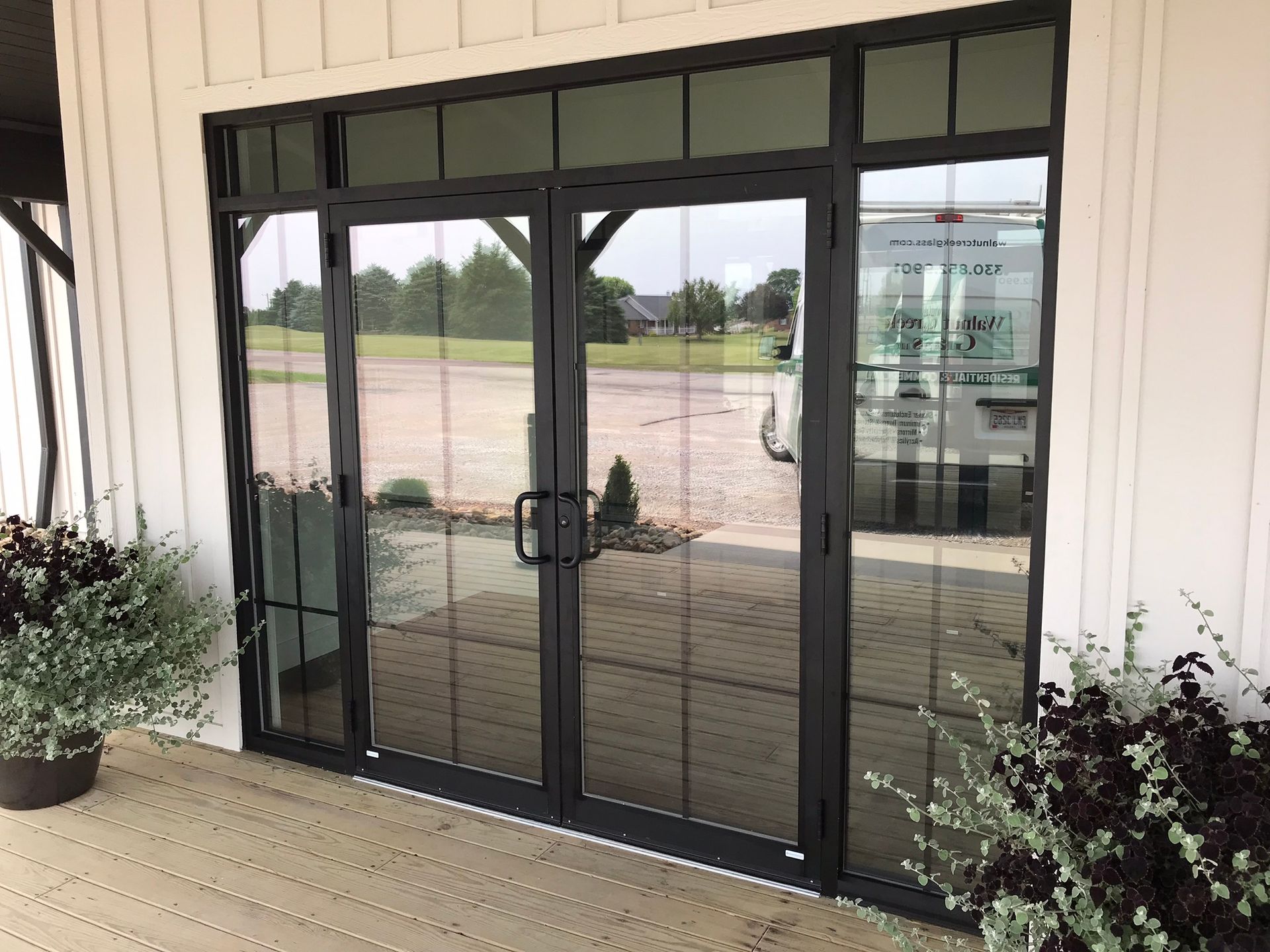 Black-framed glass doors reflecting a paved area, bordered by white siding and plants.