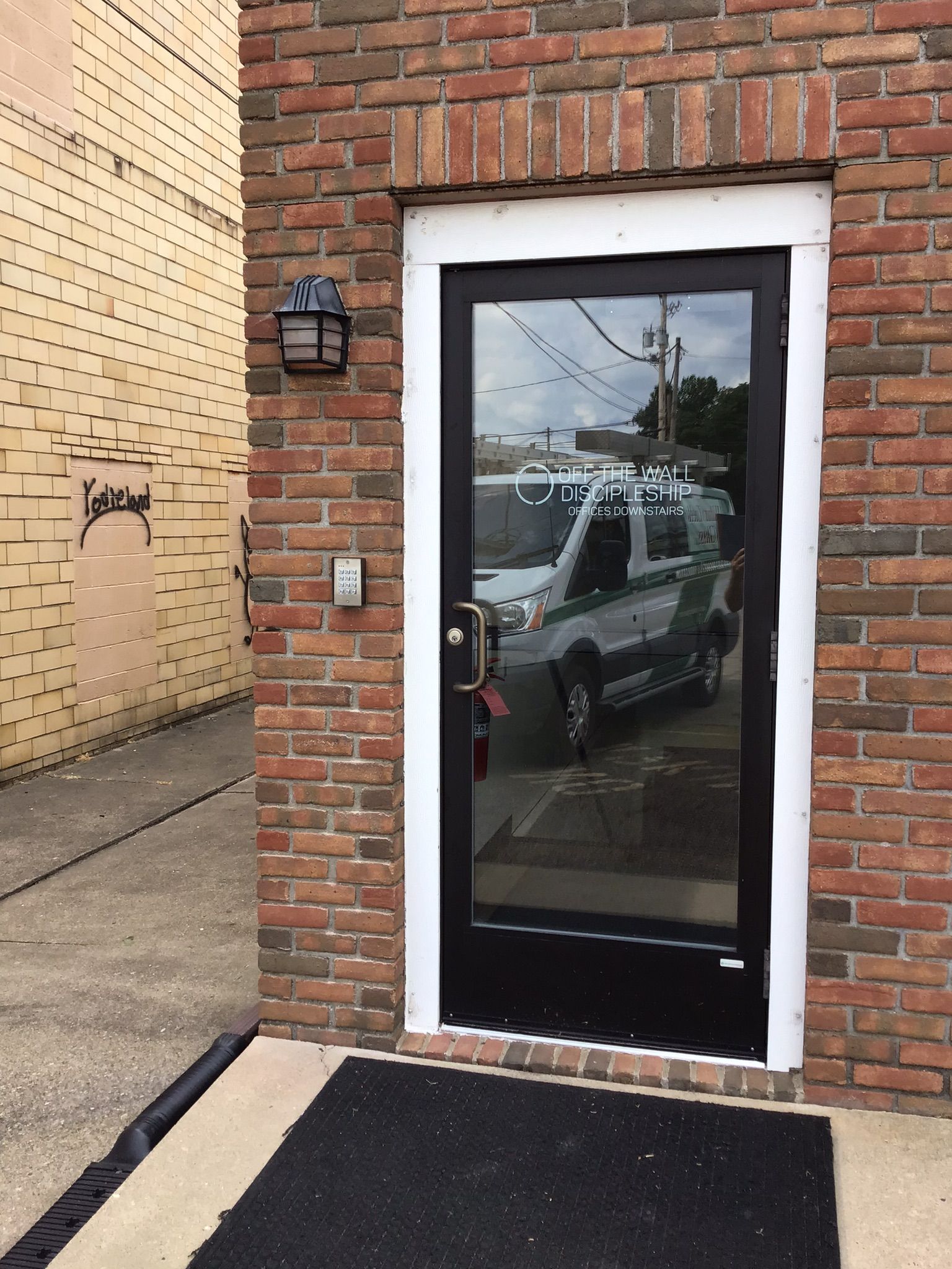 Exterior door with black frame and glass panel, set in a brick wall with white trim.  Doorway has a black welcome mat.