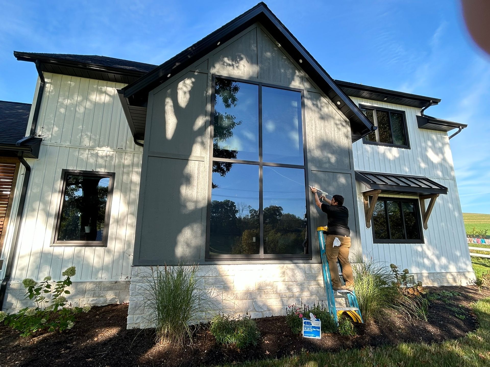 Man on ladder installing window film on a modern house with a large black-framed window.