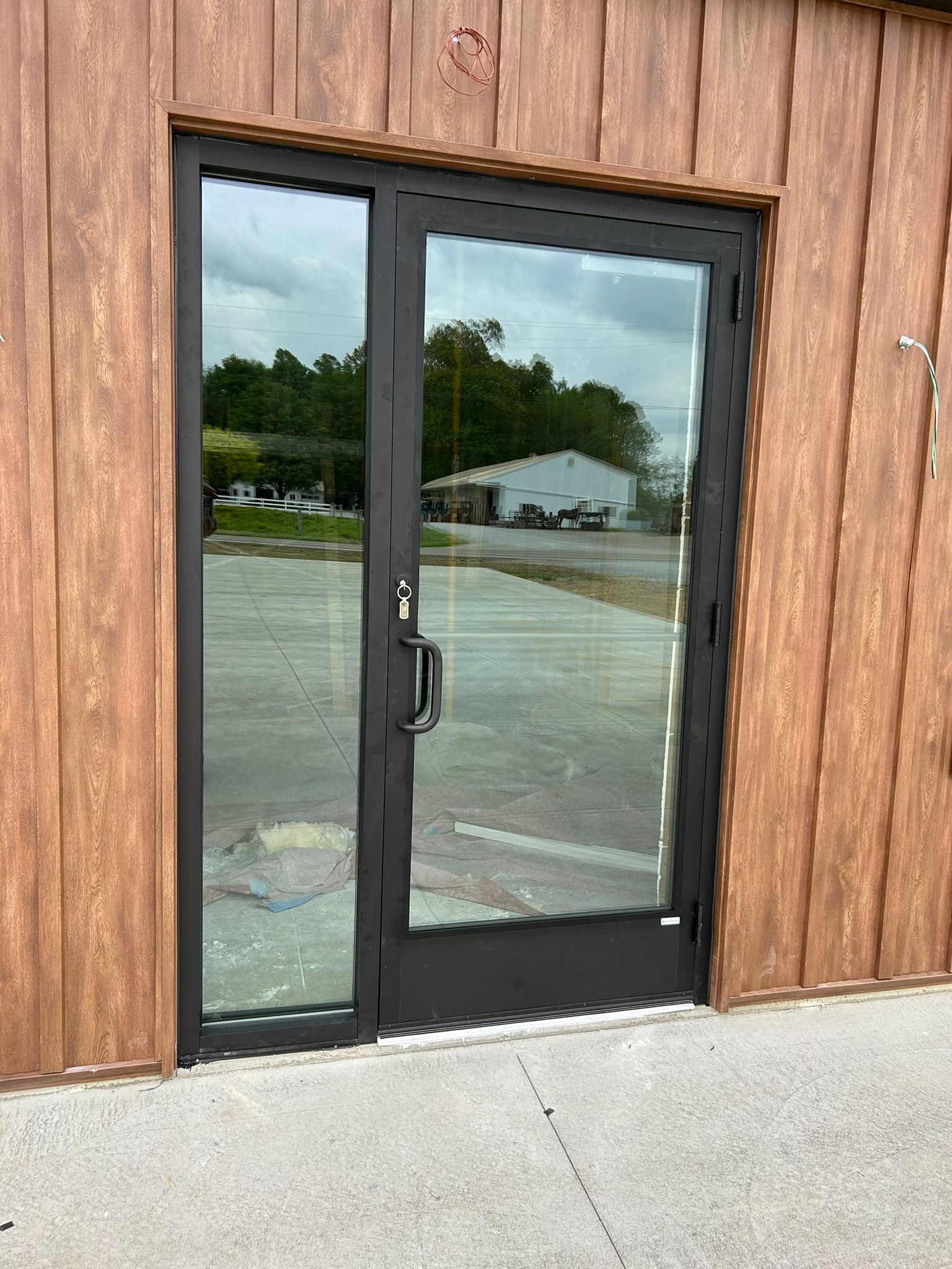 Black-framed glass door with sidelight in a brown wood-paneled building, reflecting the outside.