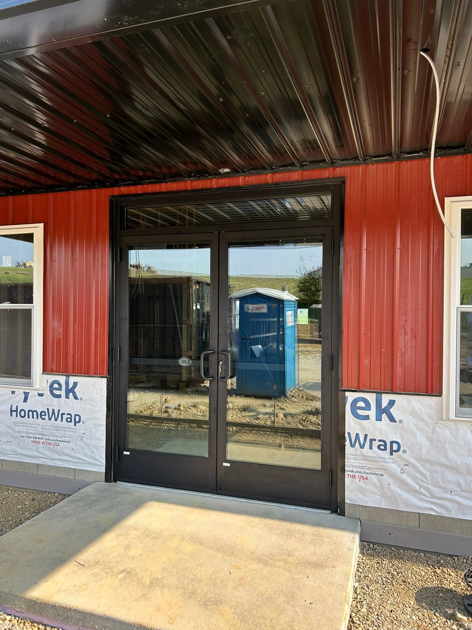 Double glass doors with black frames in a red building; a blue portable toilet is reflected in the glass.