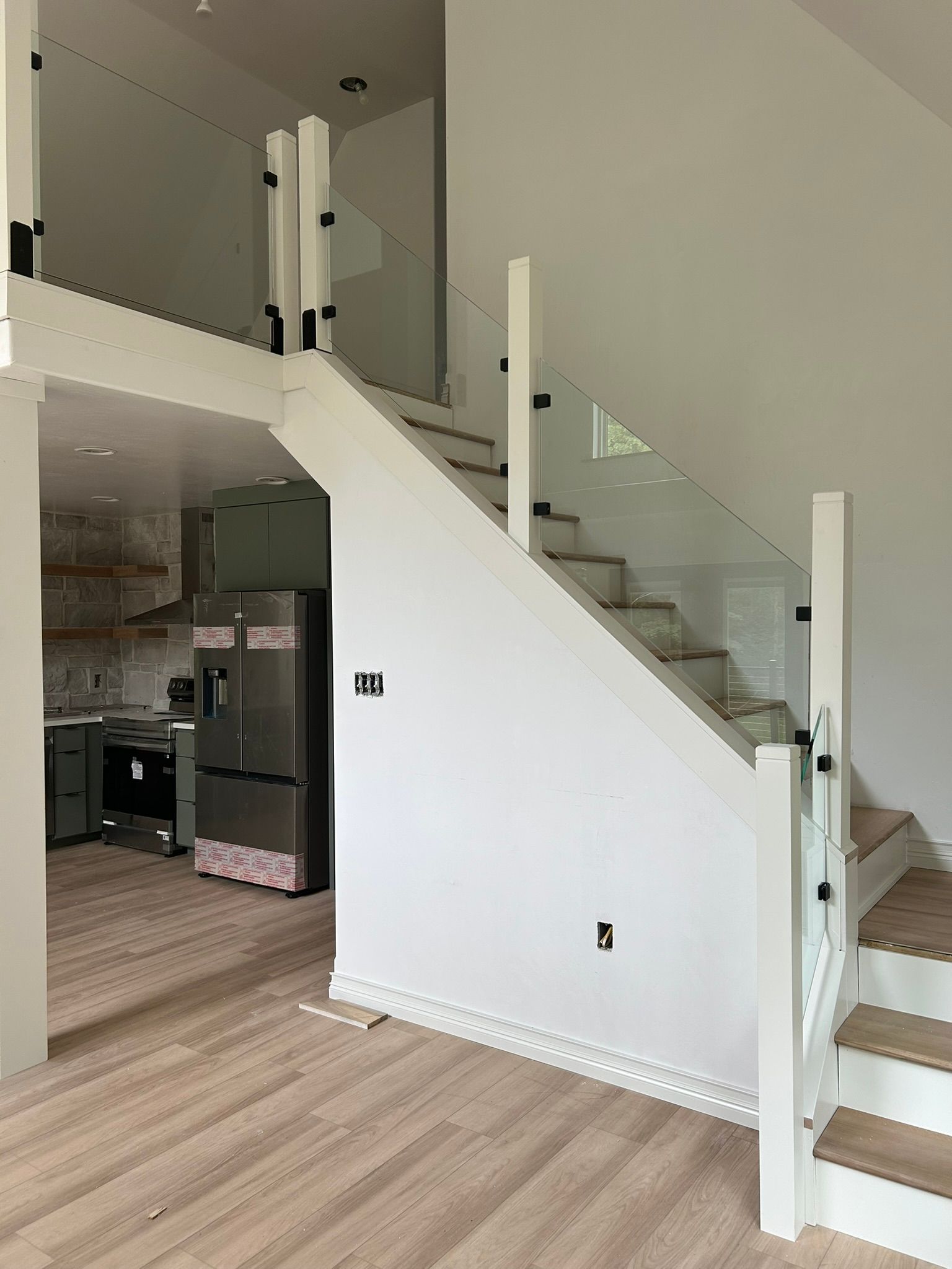 Interior view of white staircase with glass panels, hardwood floors, and a glimpse of a kitchen.