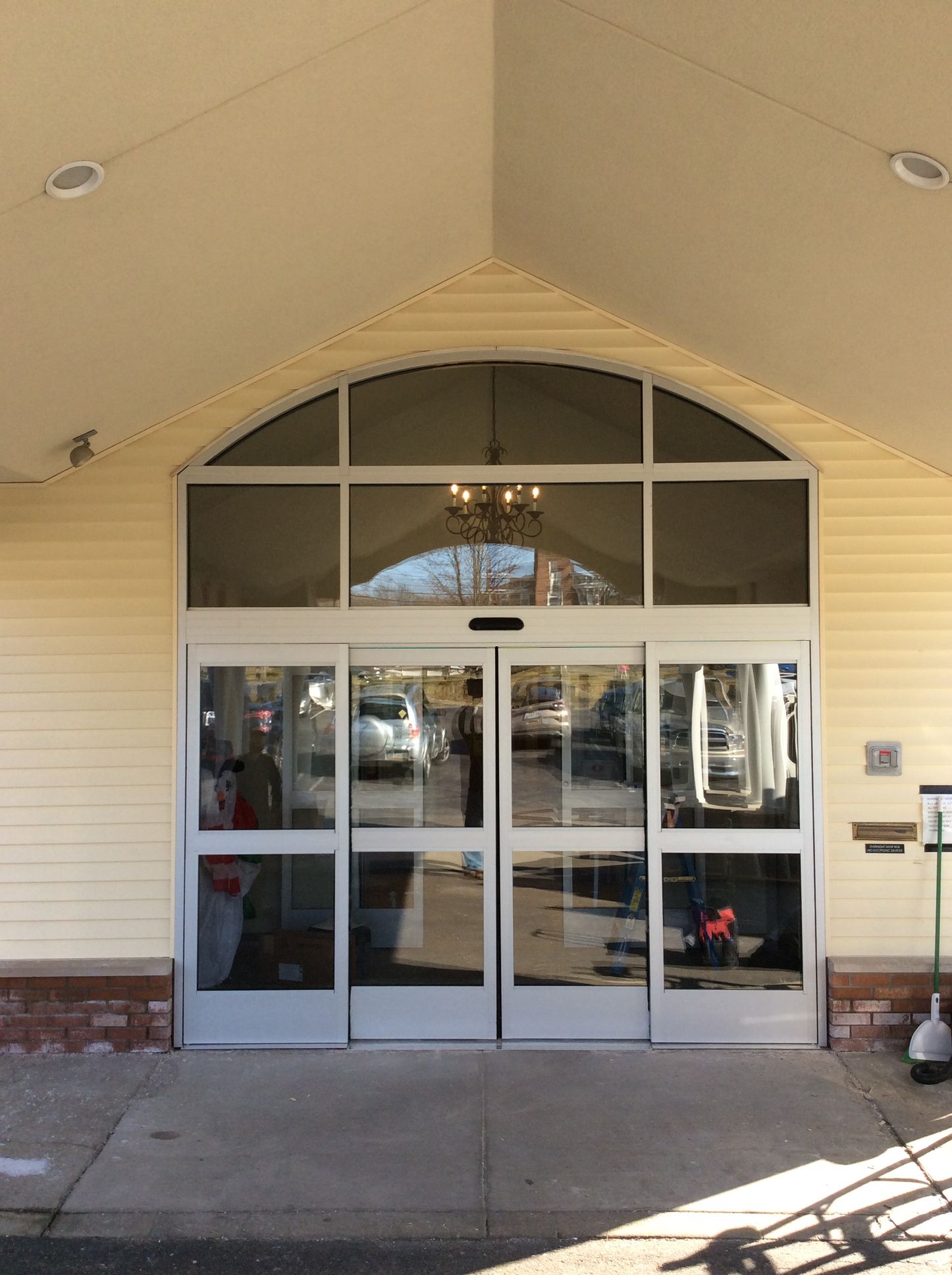 Entrance with automatic sliding doors under a beige canopy. Glass panels and a cement walkway are visible.