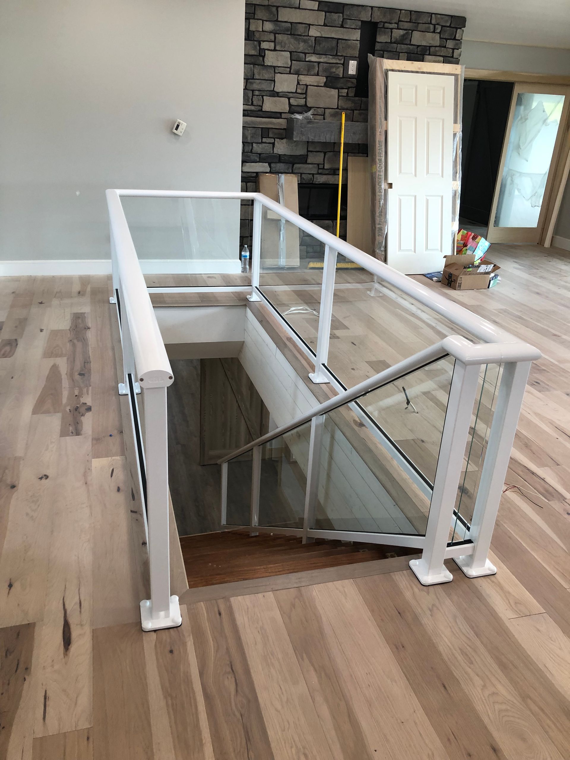 White railing and glass panels surround a staircase leading down; wood floors and a stone wall in the background.