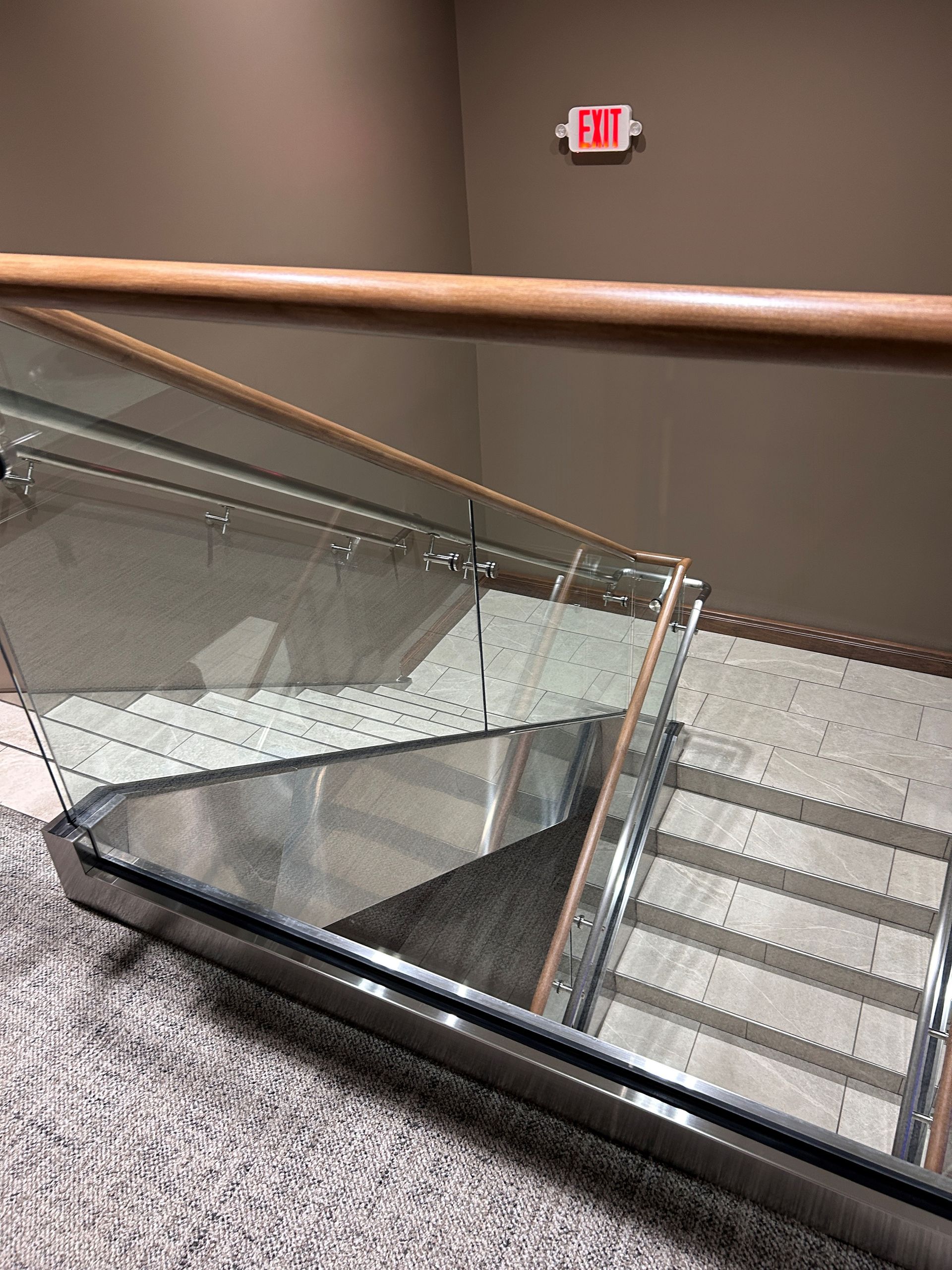 Staircase with glass railing, wood handrail, and exit sign. Gray carpeted steps and brown walls.