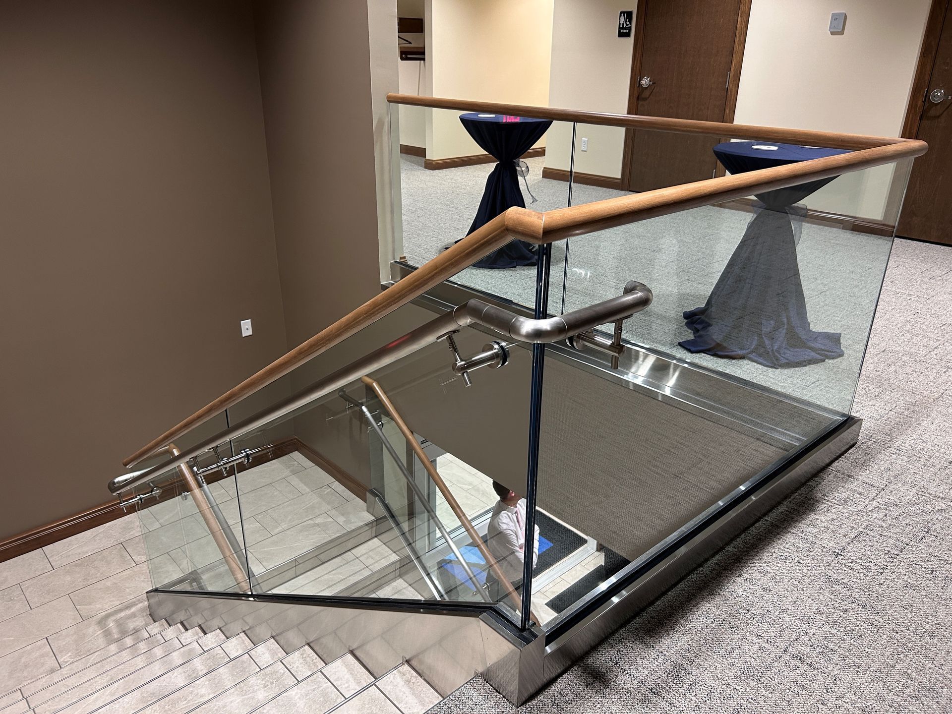 Staircase with glass and wood railings, descending to a carpeted area with tables.