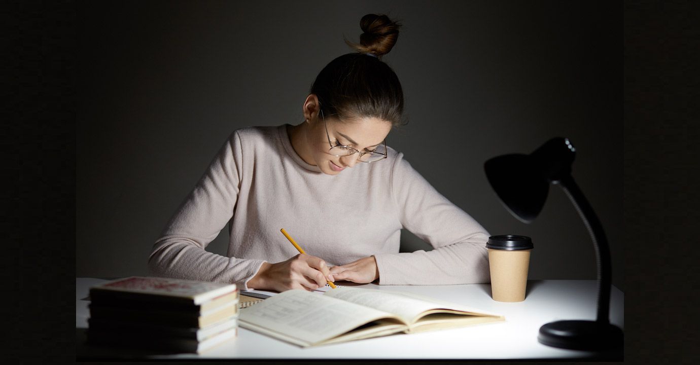 A woman is sitting at a desk with a book and a lamp.