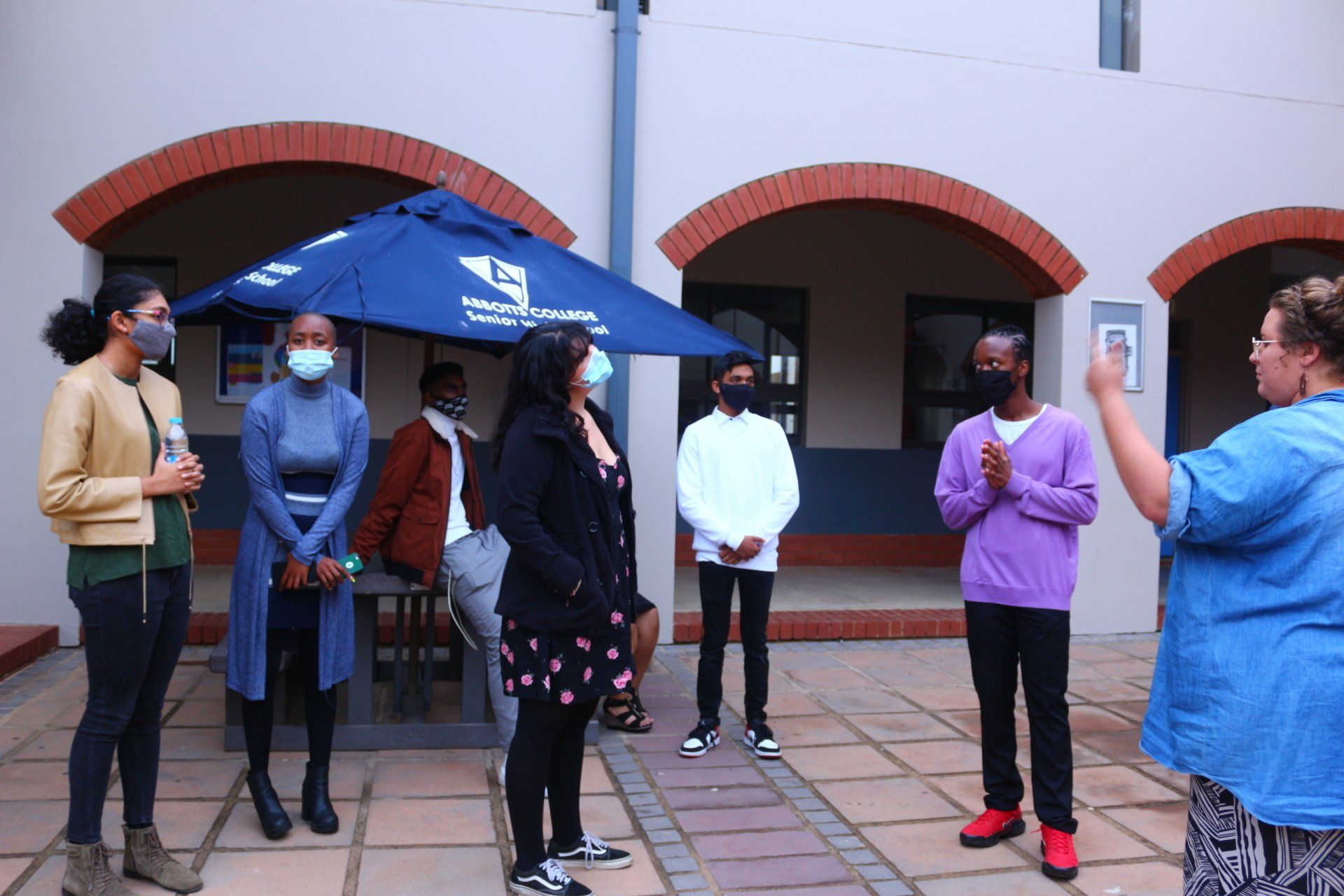 A group of people wearing masks are standing in front of a building under an umbrella.