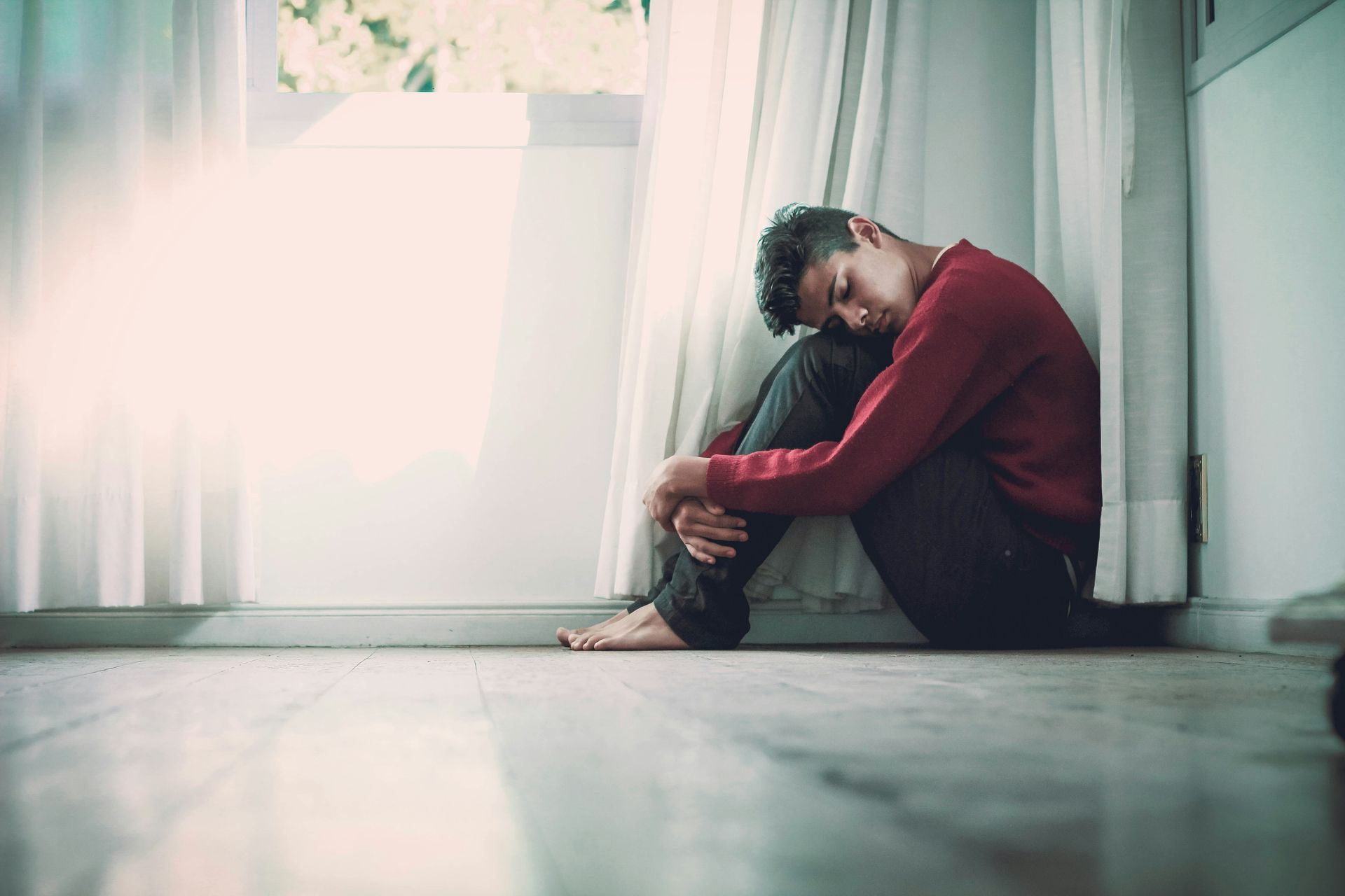 A person sits on the floor by a window, curled up with their head down, wearing a red long-sleeved shirt and dark pants.