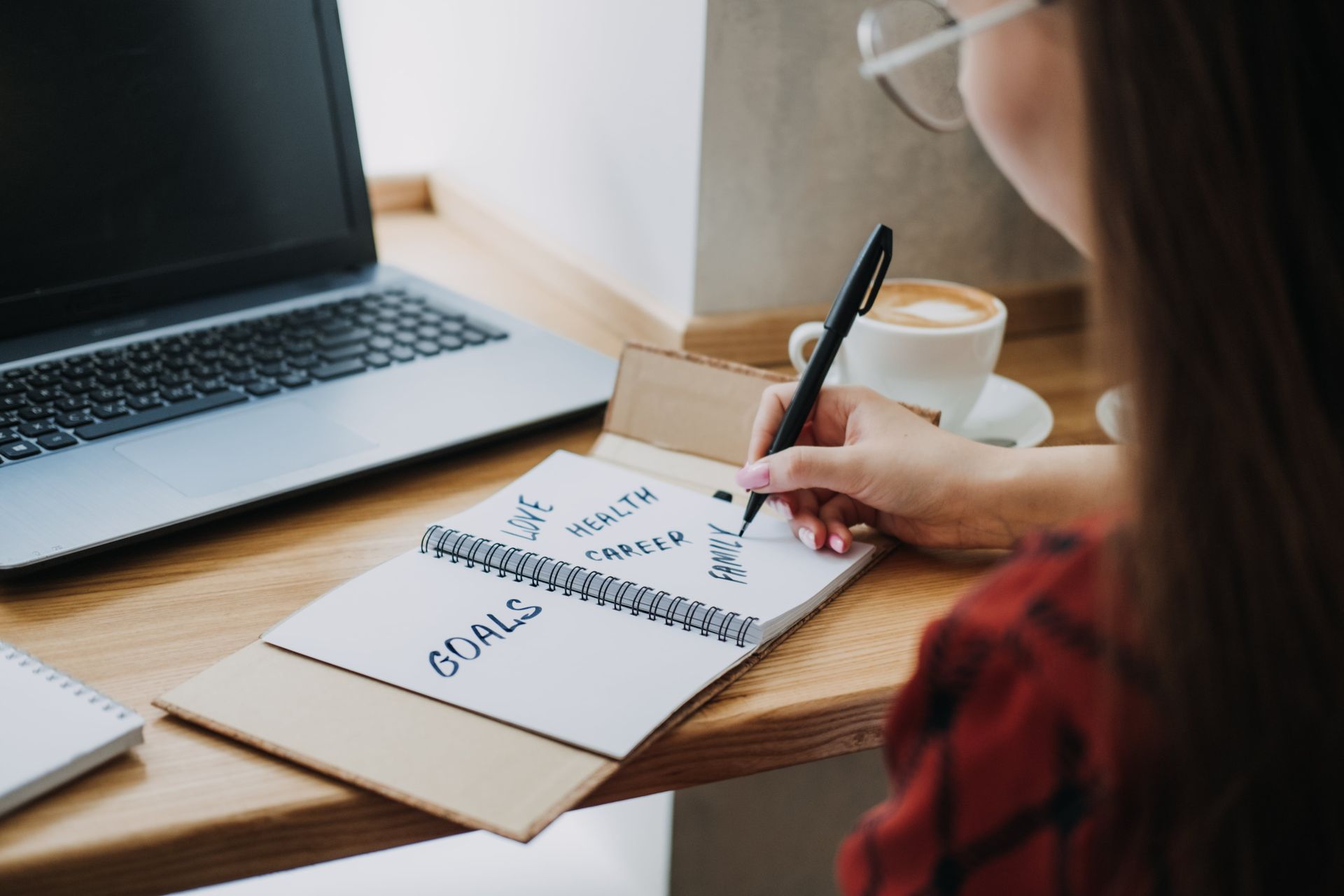 A woman is writing in a notebook while sitting at a desk with a laptop and a cup of coffee.