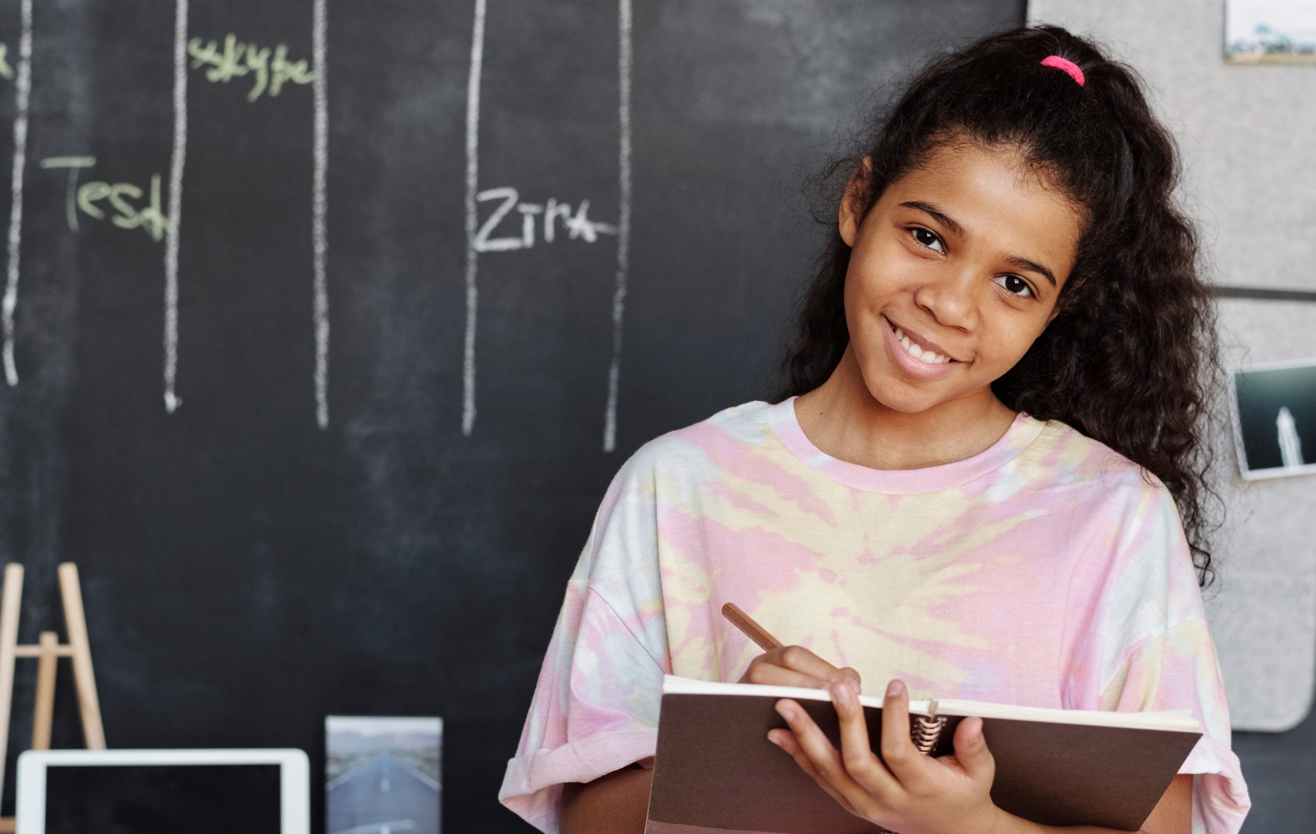 A young girl is standing in front of a blackboard holding a notebook and a pen.