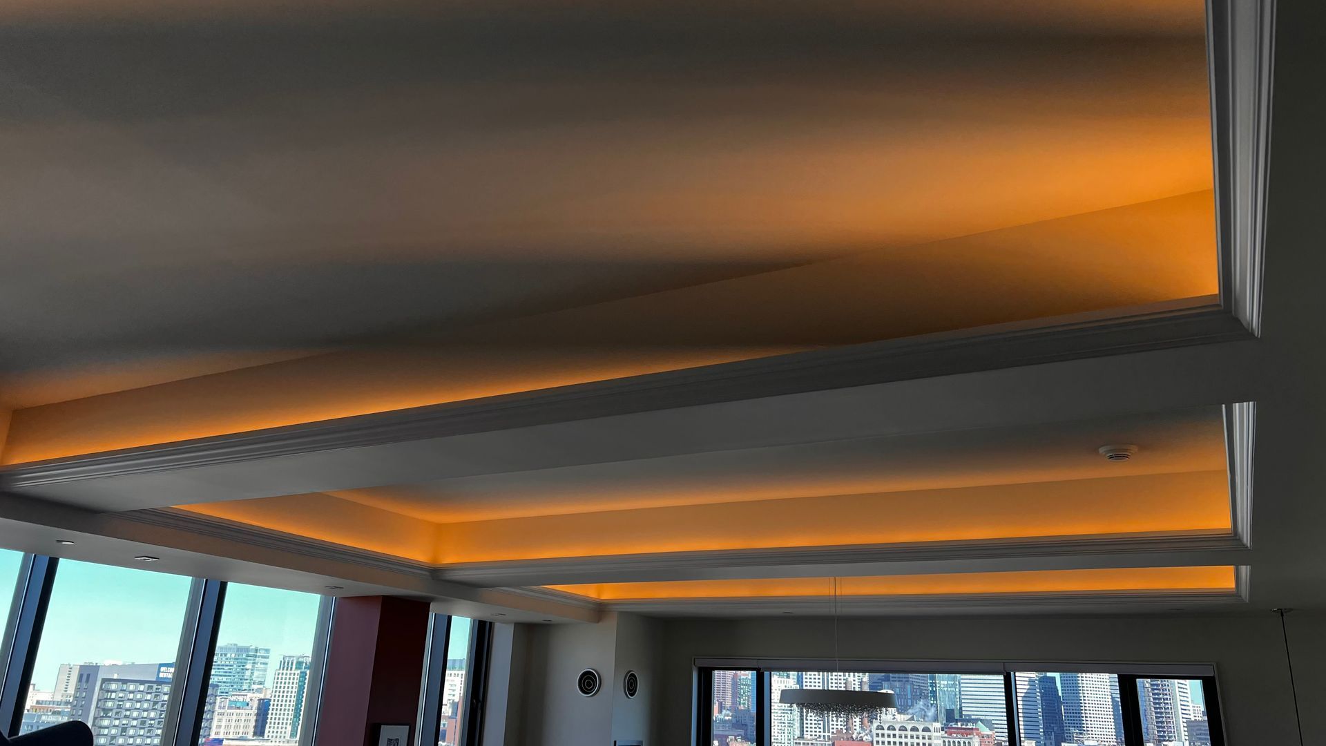 White ceiling with recessed lighting glows orange; tall windows reveal a cityscape.