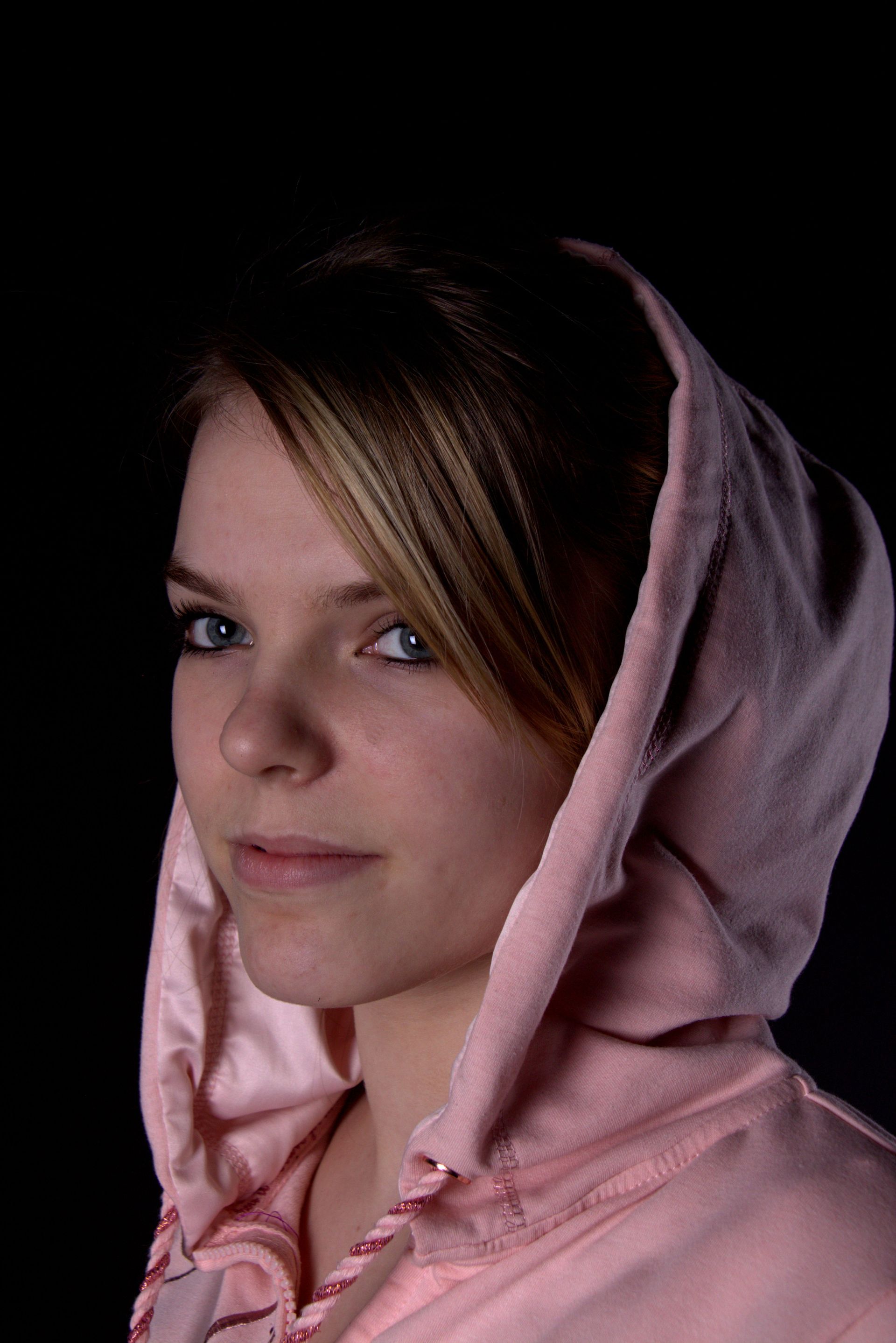 Portret van een persoon in een roze hoodie tegen een zwarte achtergrond, die in de camera kijkt.