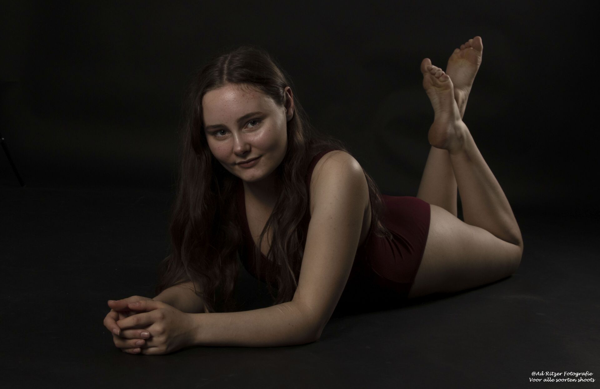 Vrouw poseert op haar buik in een donkere studio, gekleed in een bordeauxrode outfit en lachend naar de camera.