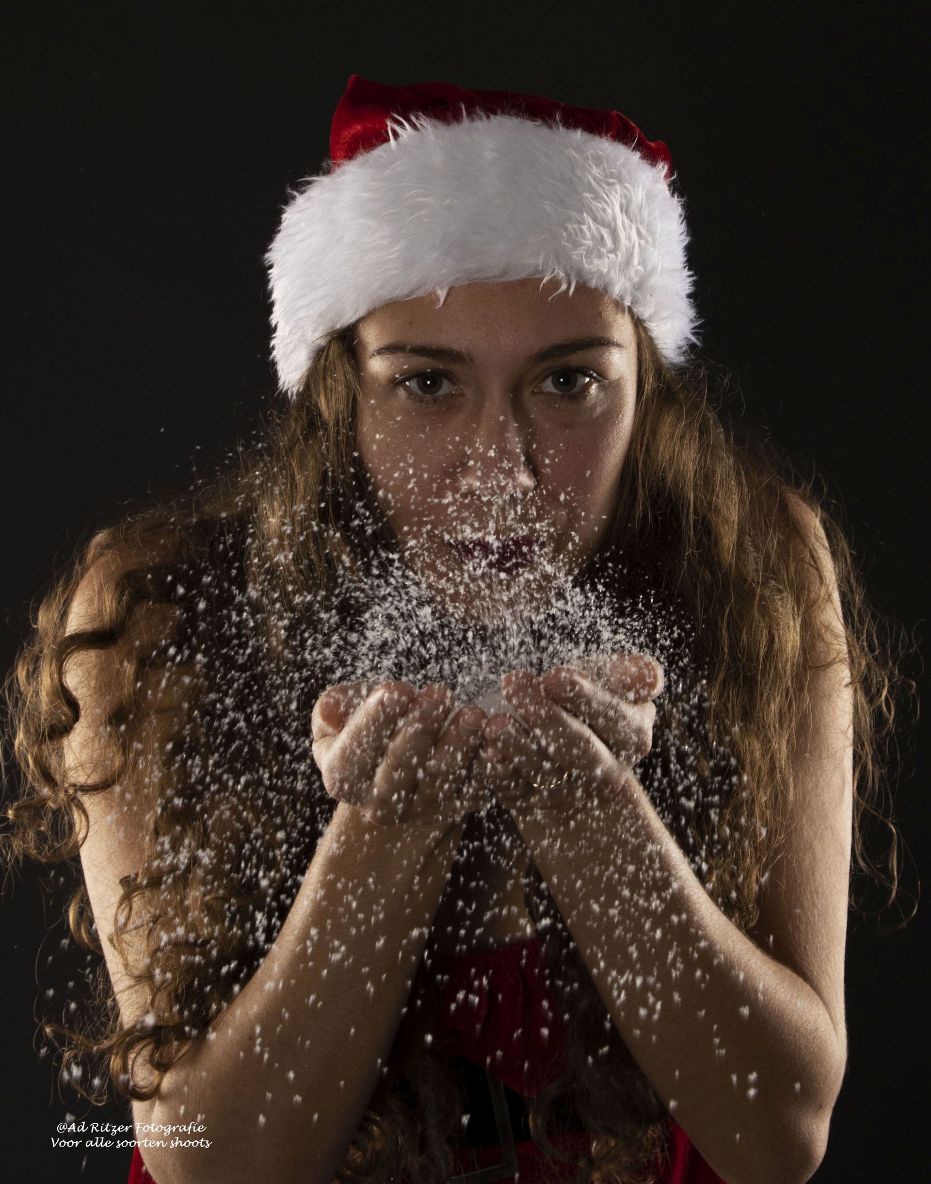 Vrouw met een kerstmuts blaast glitter uit haar handen tegen een donkere achtergrond.