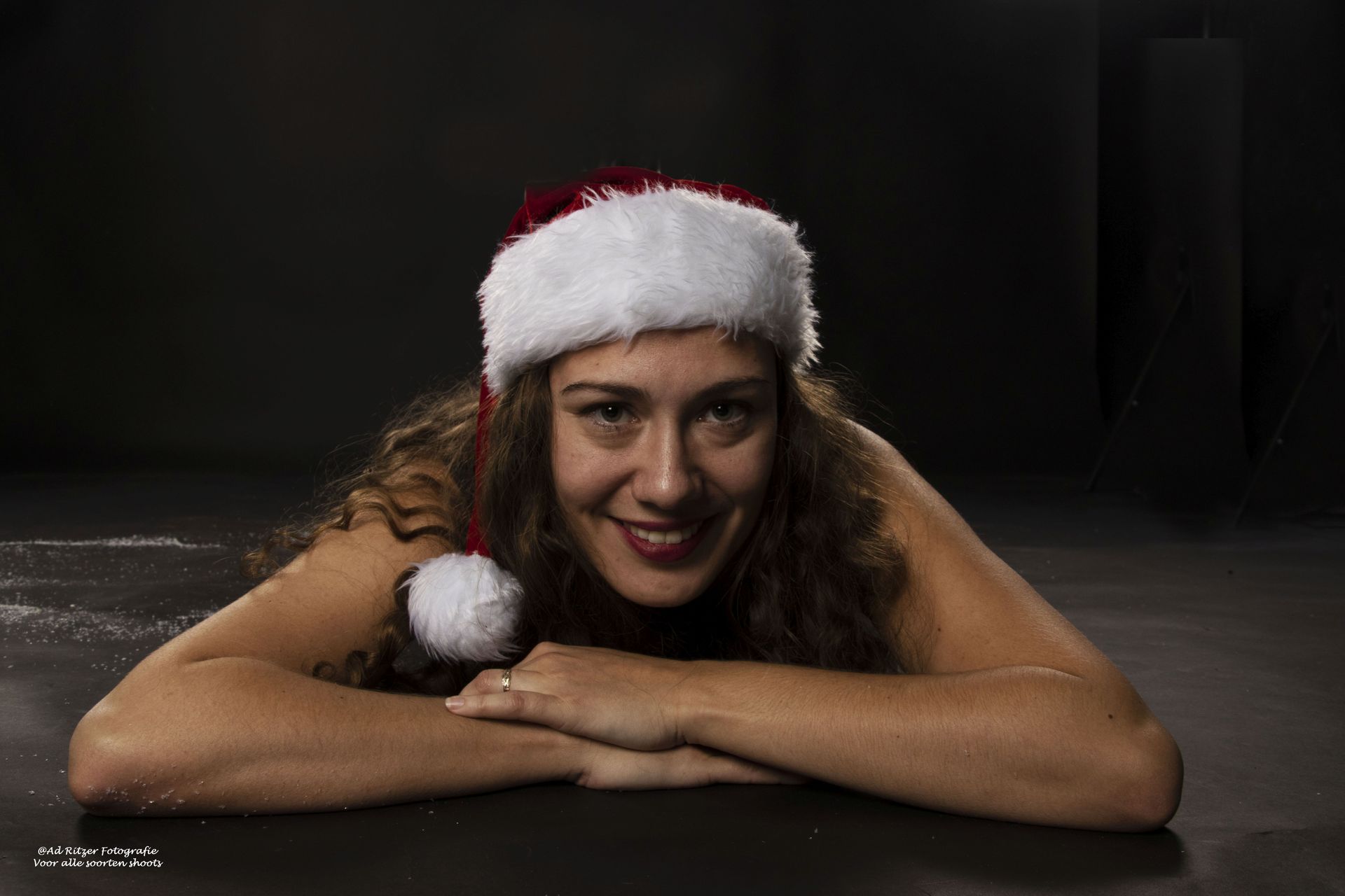 Vrouw met een kerstmuts op, lachend met haar kin op haar gevouwen armen, tegen een donkere achtergrond.